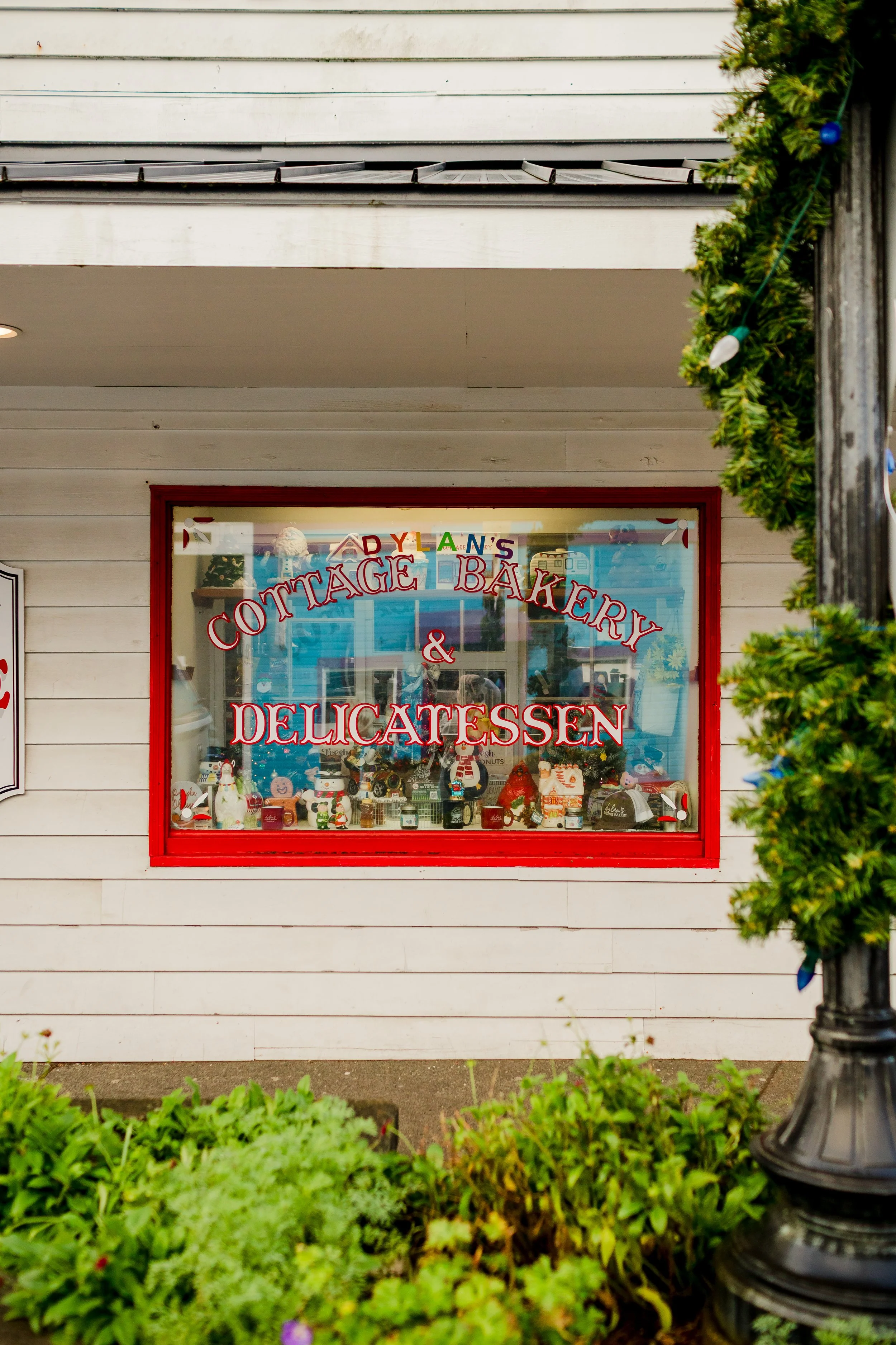 Storefront with a sign reading 'Dylan's Cottage Bakery & Delicatessen,' decorated with Christmas ornaments, under a balcony and next to a green bush.