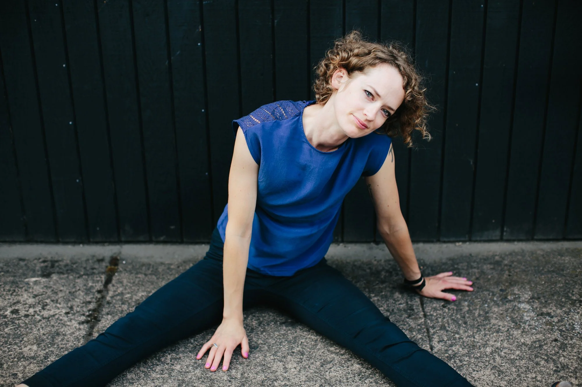 A woman with curly hair sitting on the ground in a split position against a black wooden wall, wearing a blue shirt and black pants.