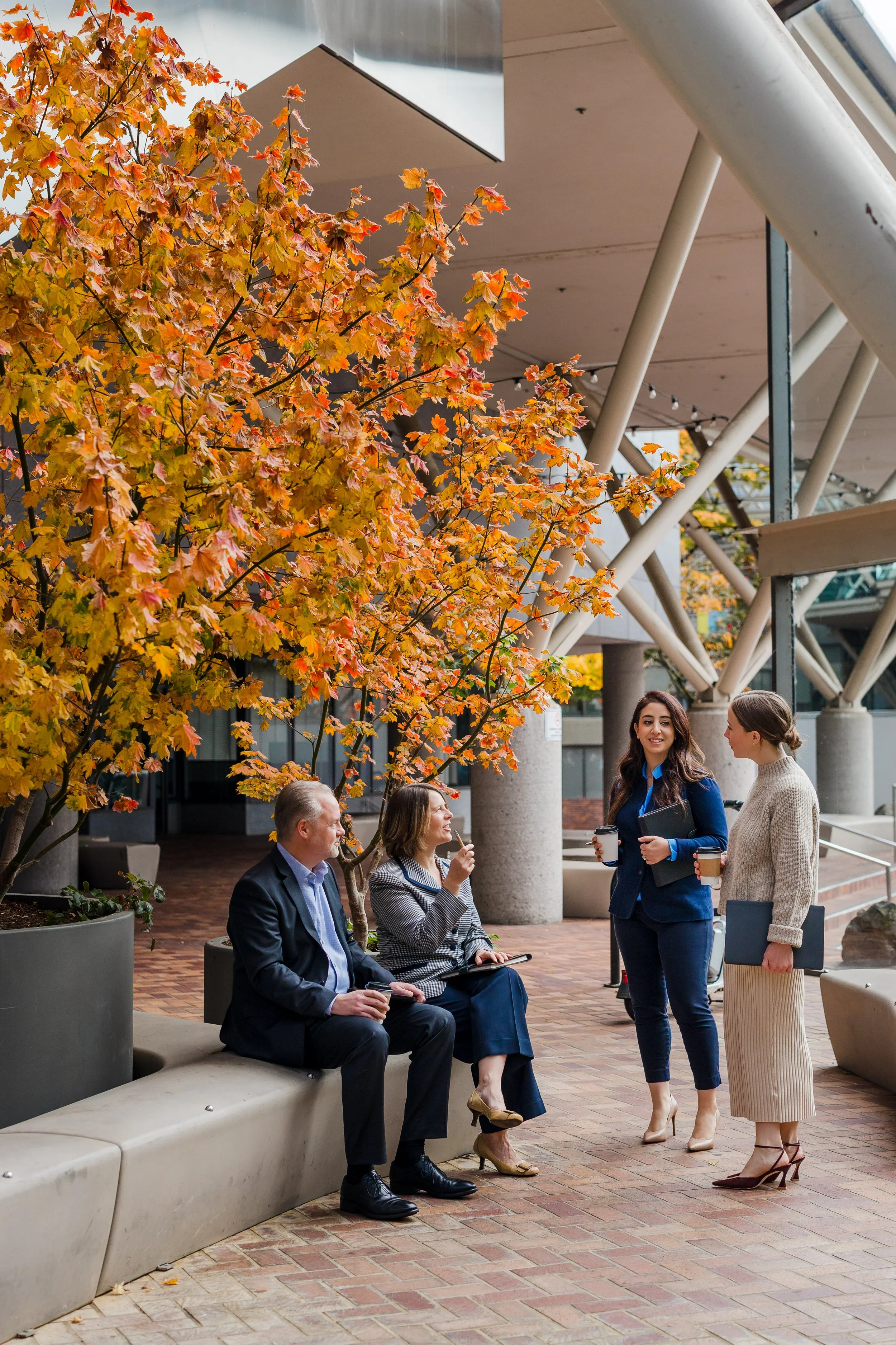 Four businesspeople, two seated and two standing, engaging in a lively conversation outdoors under a tree with colorful autumn leaves, near a modern building with large windows and concrete pillars.