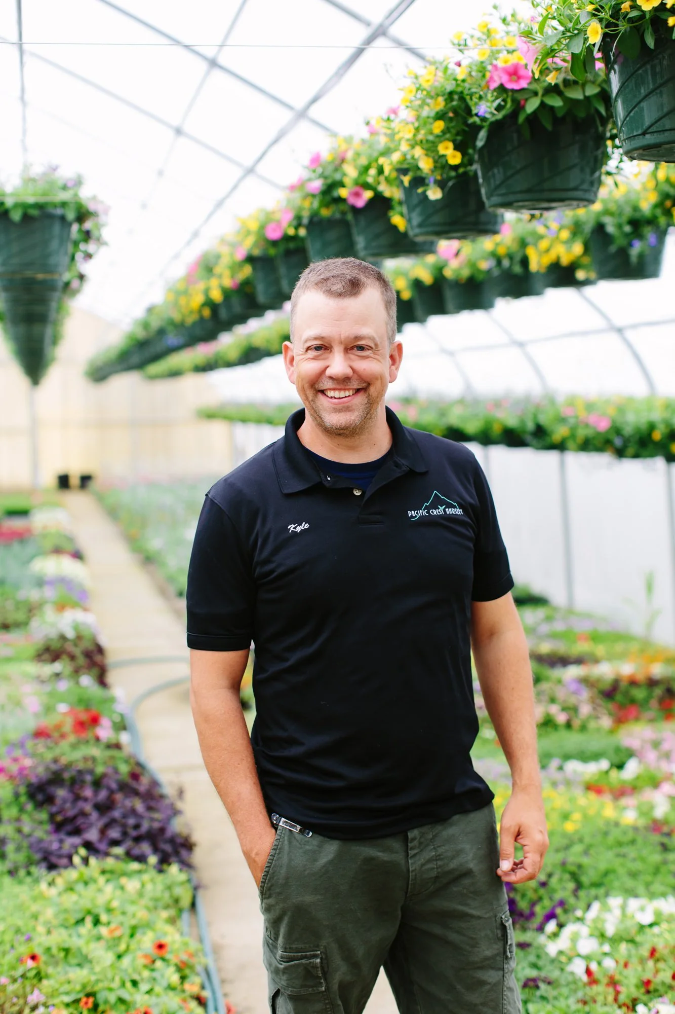 A man smiling and standing inside a greenhouse with hanging flower pots containing colorful flowers.