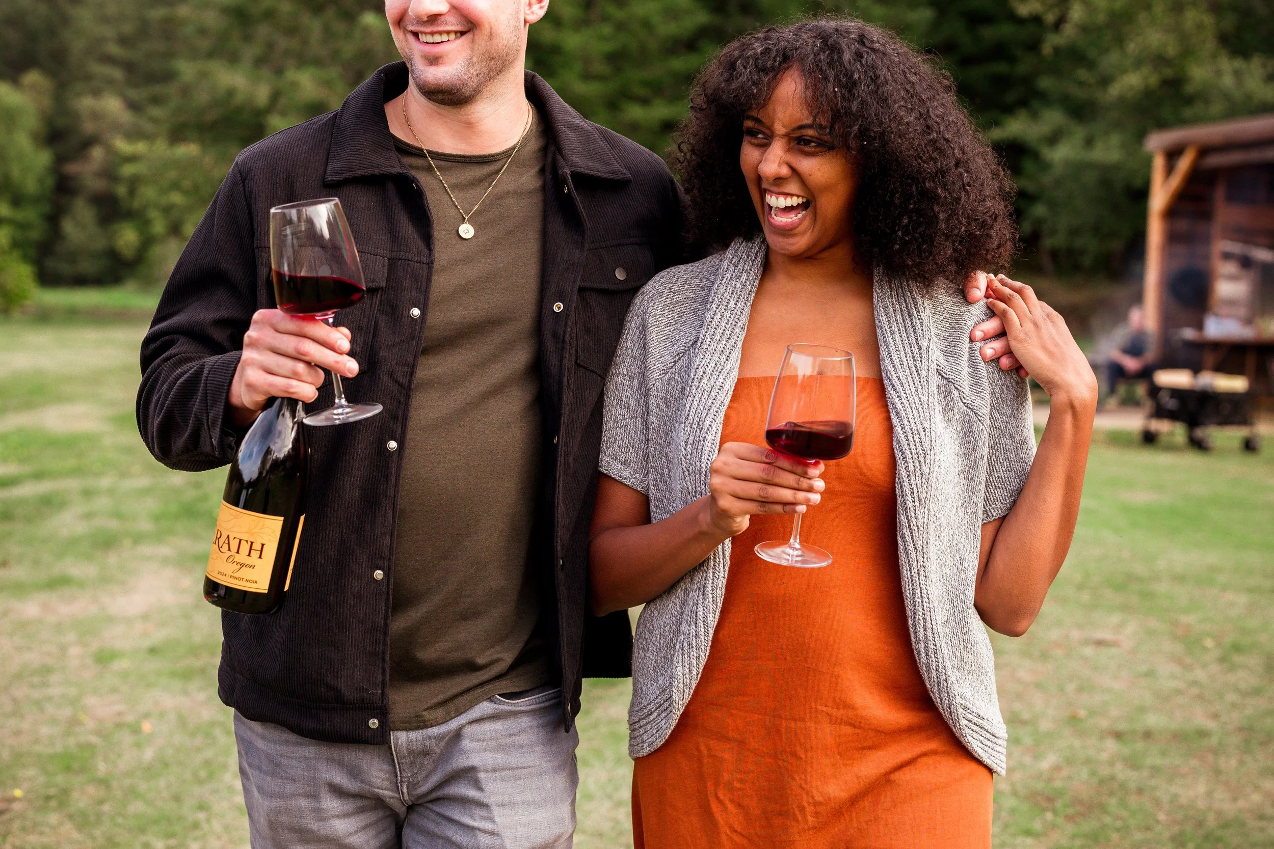 A man and woman outdoors holding glasses of red wine, smiling and enjoying each other's company at a park or garden setting.