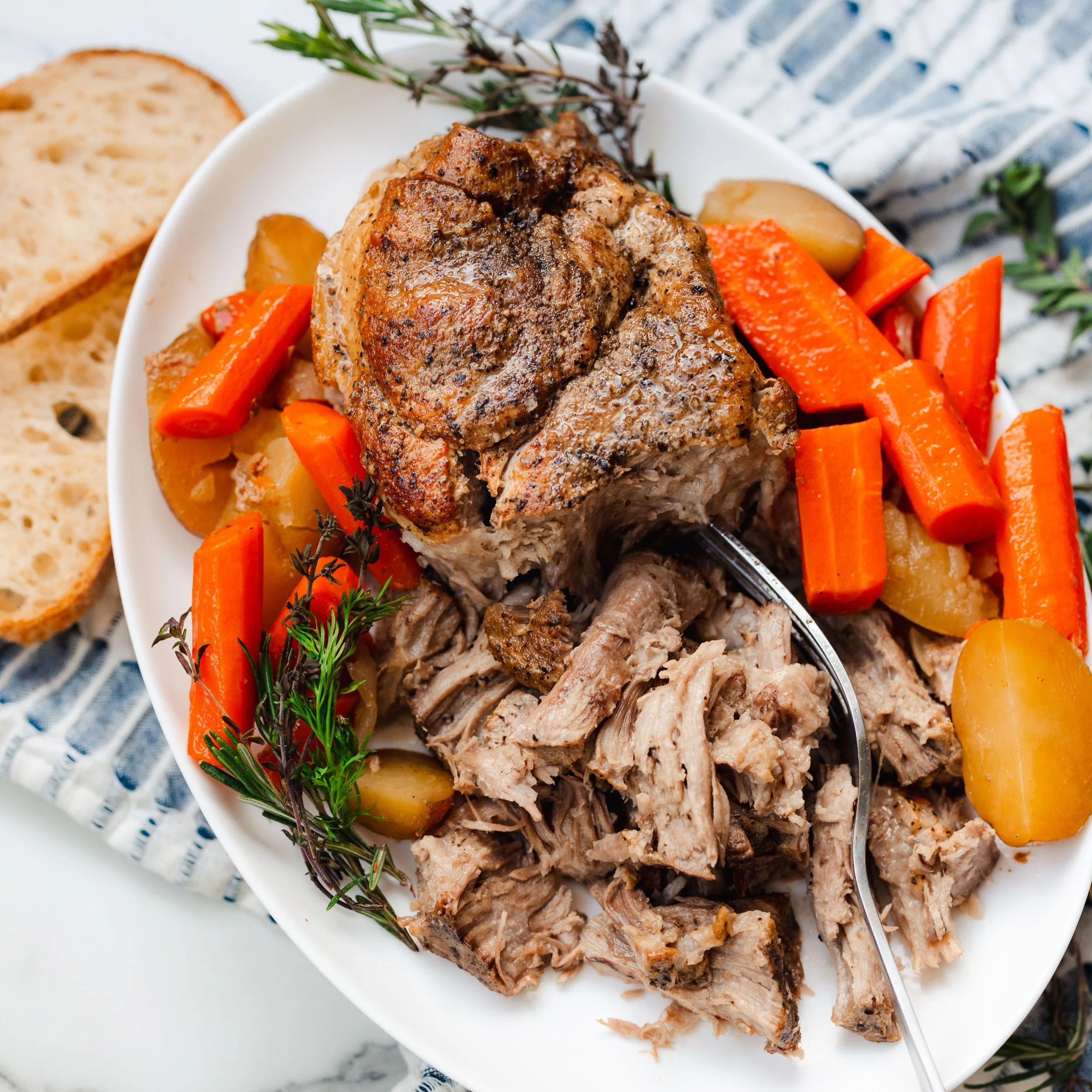 Roast pork with carrots and potatoes on a white plate, garnished with fresh herbs and slices of bread nearby.