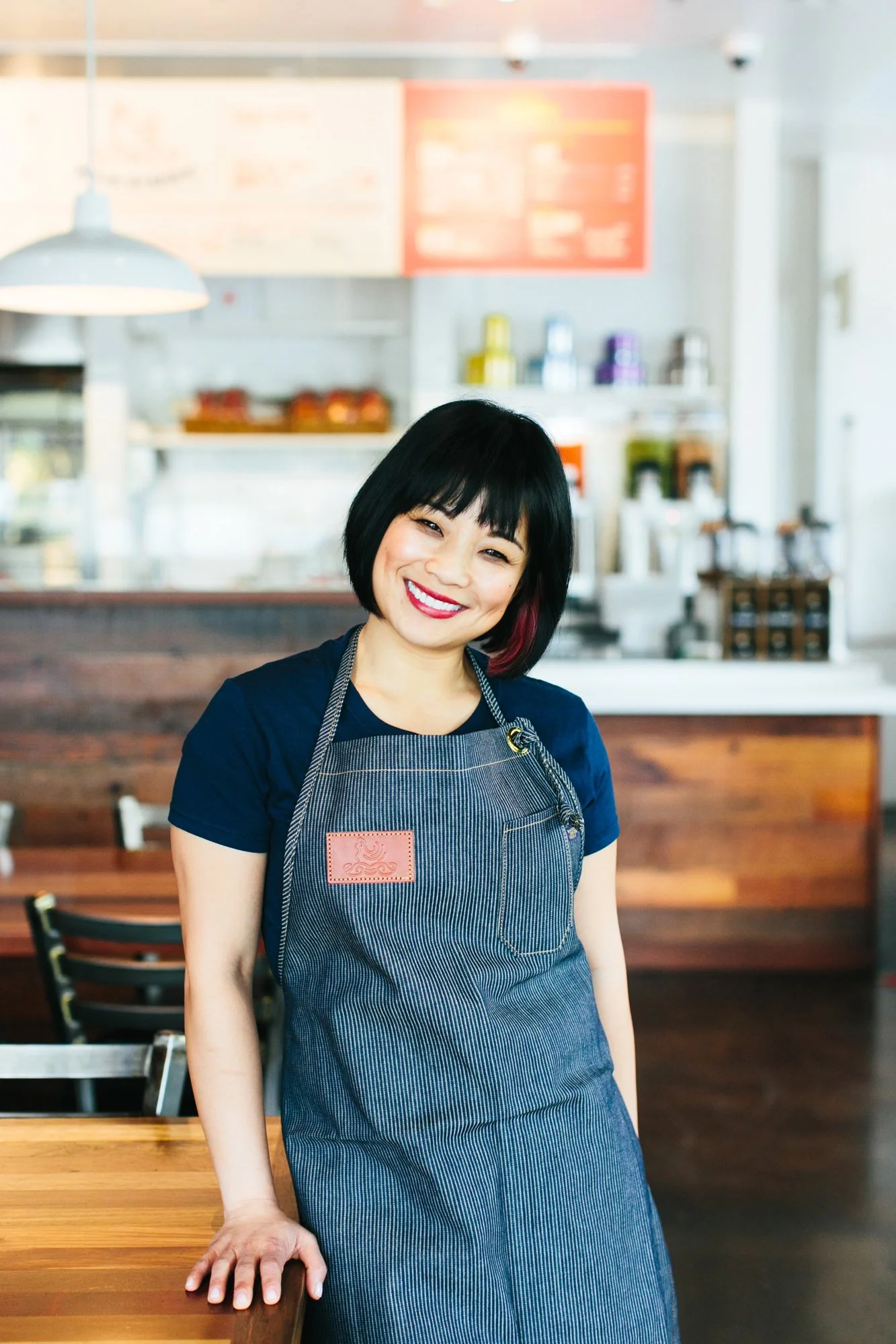 A smiling Asian woman with short black hair and red lipstick, wearing a dark blue shirt and a striped apron, stands in a cafe or restaurant.