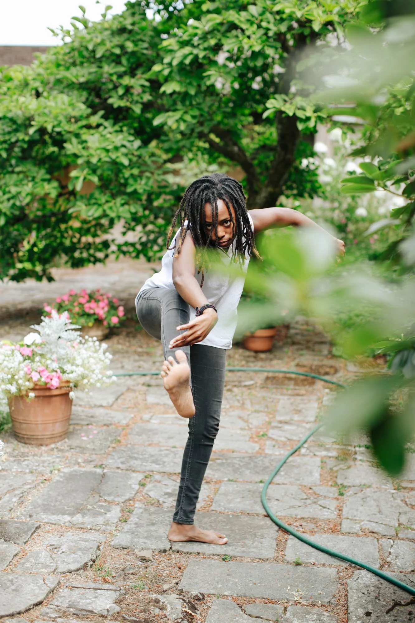 Person with dreadlocks practicing yoga outdoors on a stone pathway, surrounded by green trees and pink and white flowers in pots.