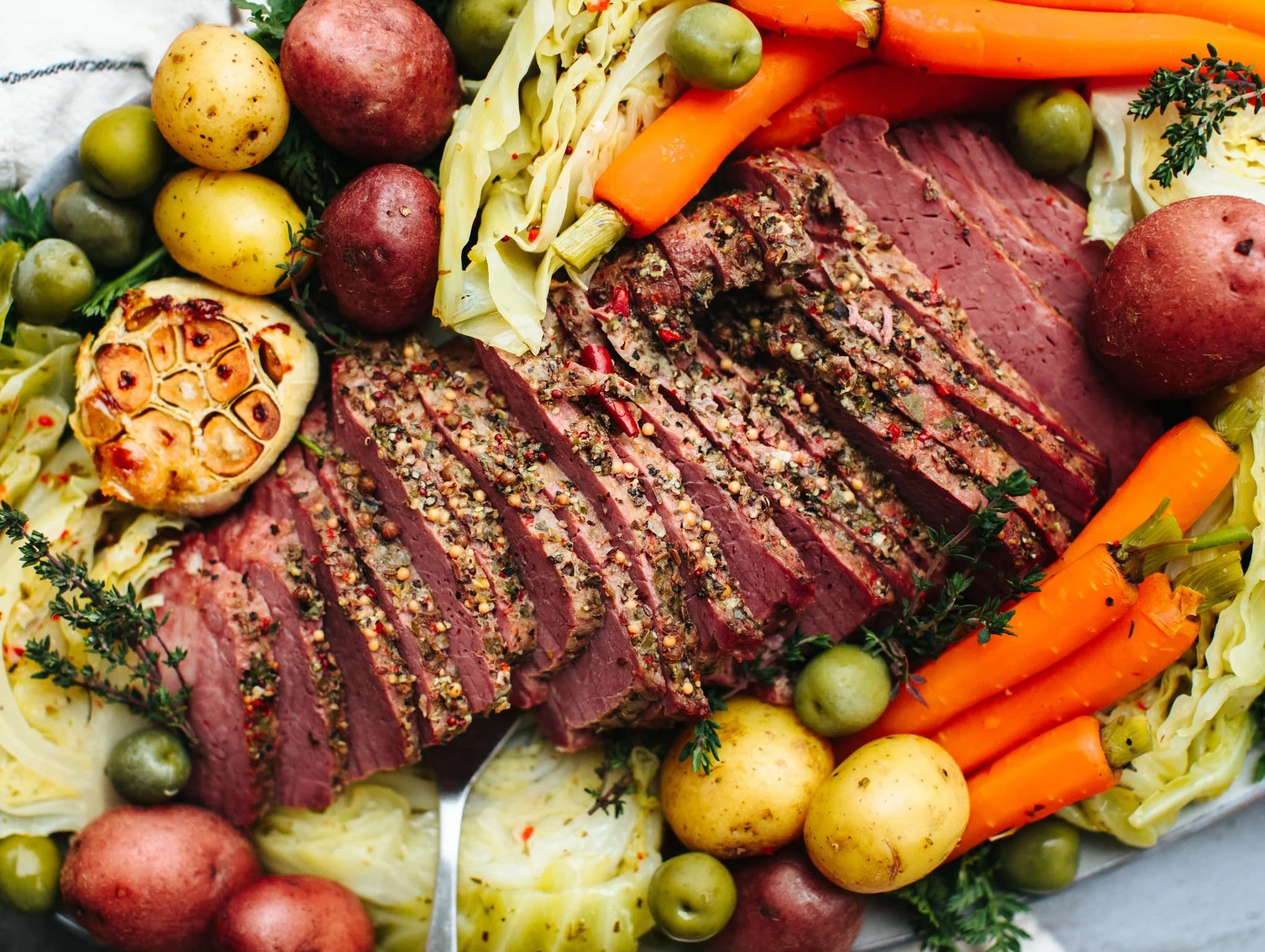 Sliced medium-rare beef tenderloin seasoned with spices, garnished with herbs, surrounded by roasted garlic, small potatoes, carrots, cabbage, and green olives.