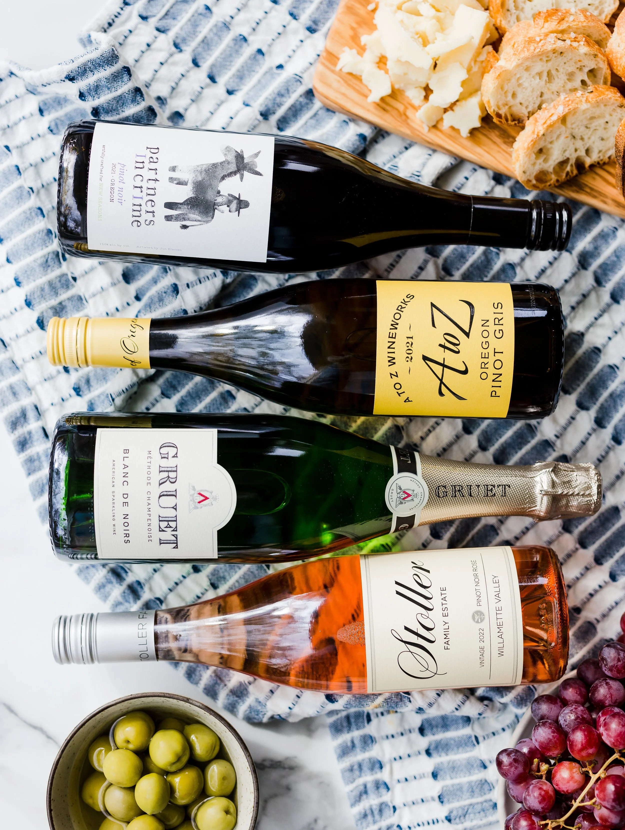 Three bottles of wine, a bowl of green olives, a bunch of red grapes, and a wooden board with cheese and bread slices on a table covered with a blue striped cloth.
