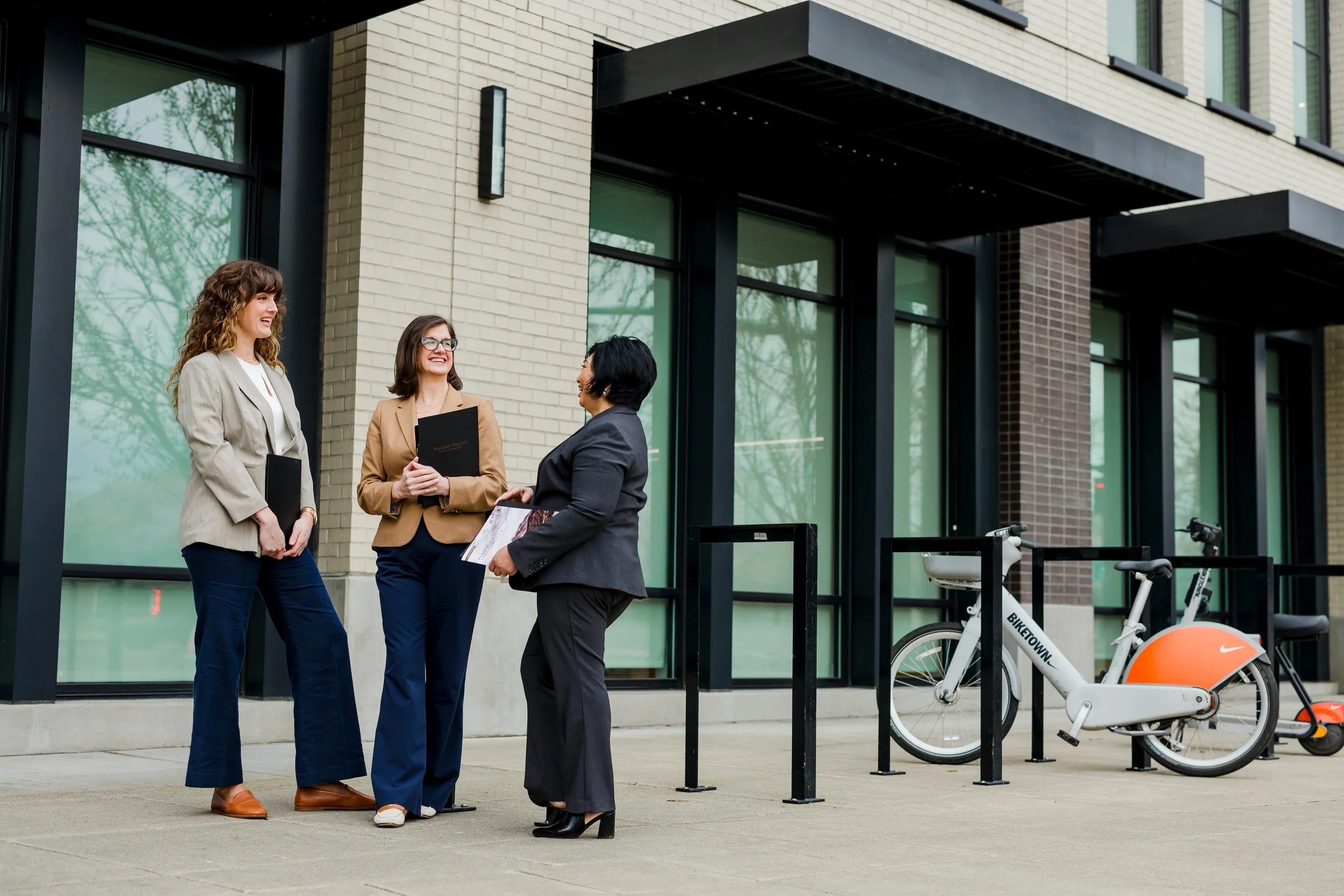 Three women in business attire standing outside a modern office building, engaged in conversation, with a shared bicycle parked nearby.