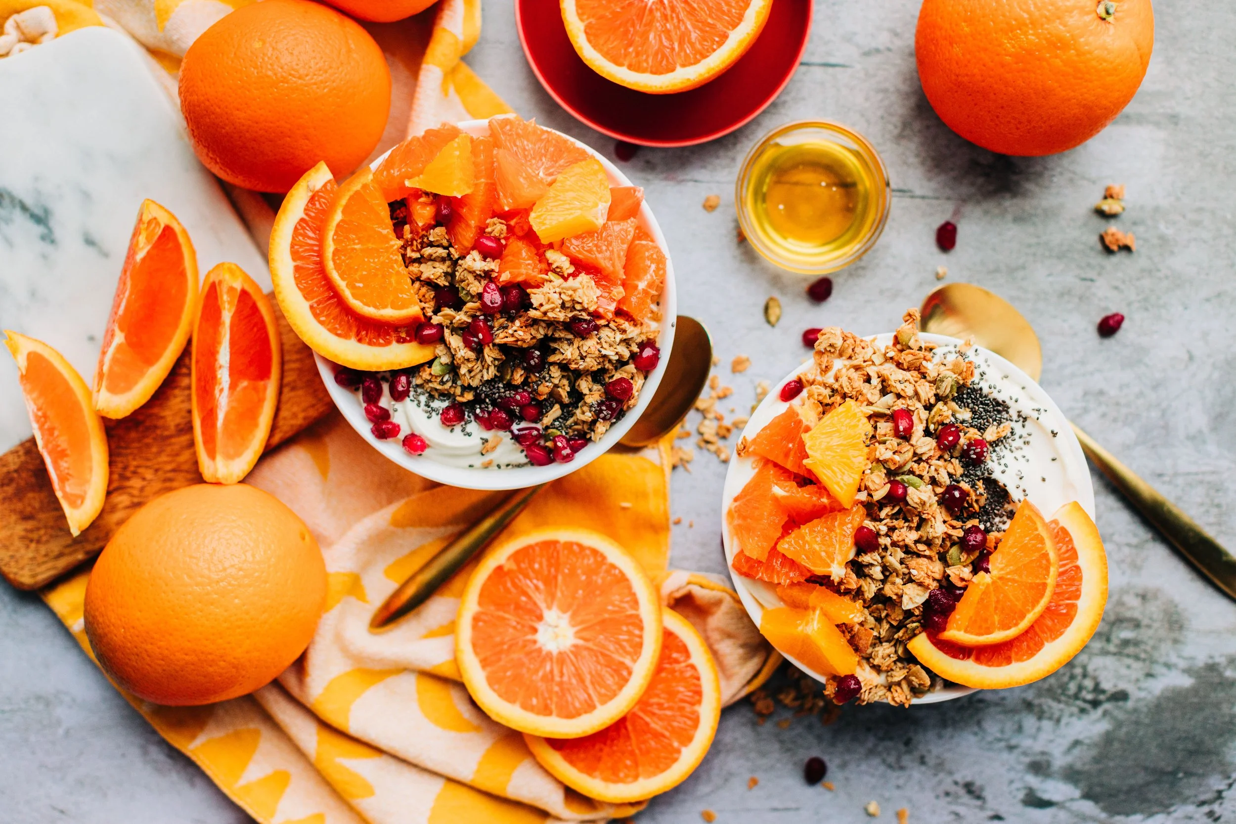 A flat lay of sliced oranges and bowls of yogurt topped with granola and orange slices, across a gray surface with scattered pomegranate seeds and honey.