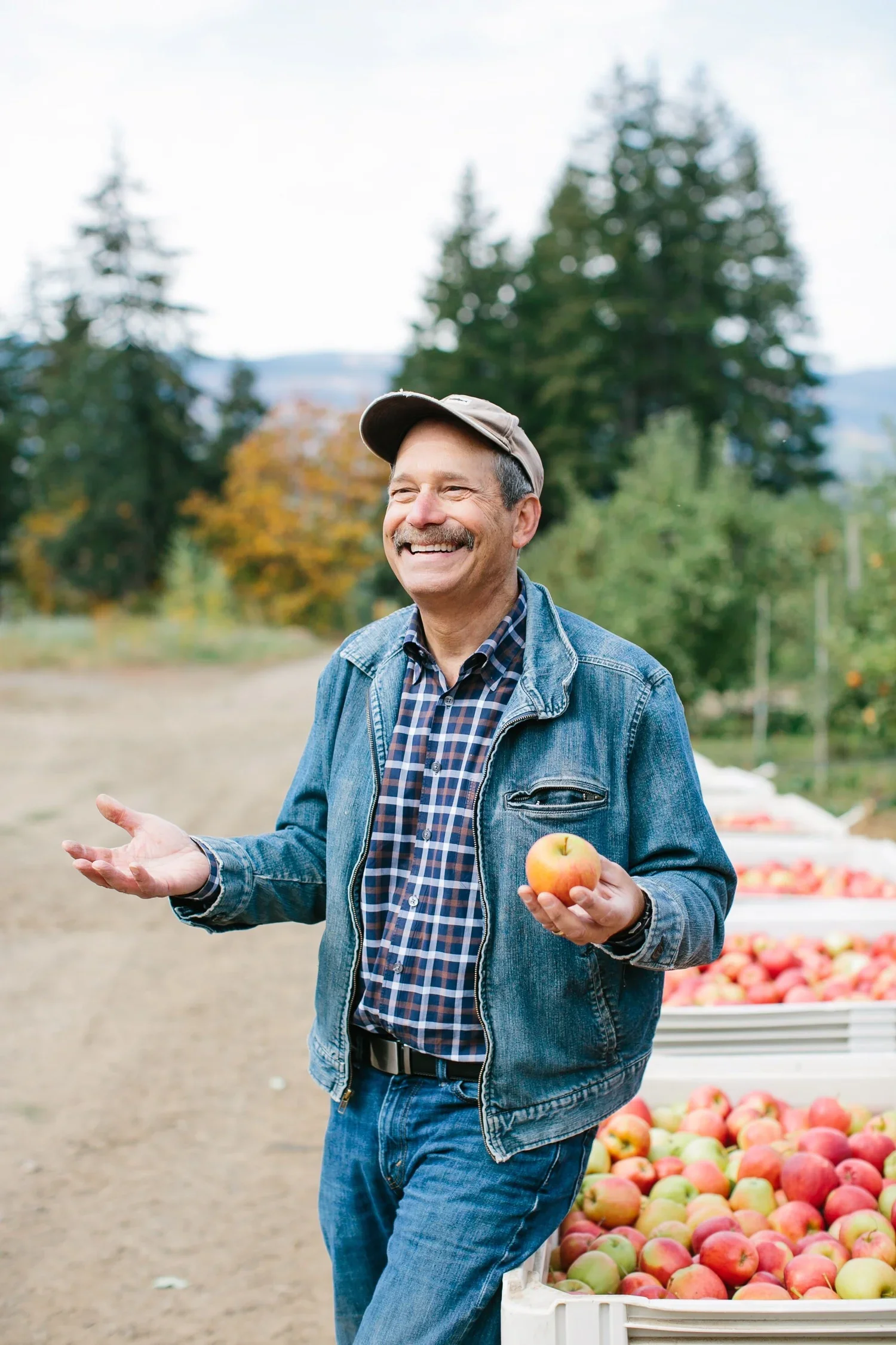 Smiling man with a mustache wearing a denim jacket, plaid shirt, and hat, holding an apple in his right hand, standing outdoors near crates of apples.