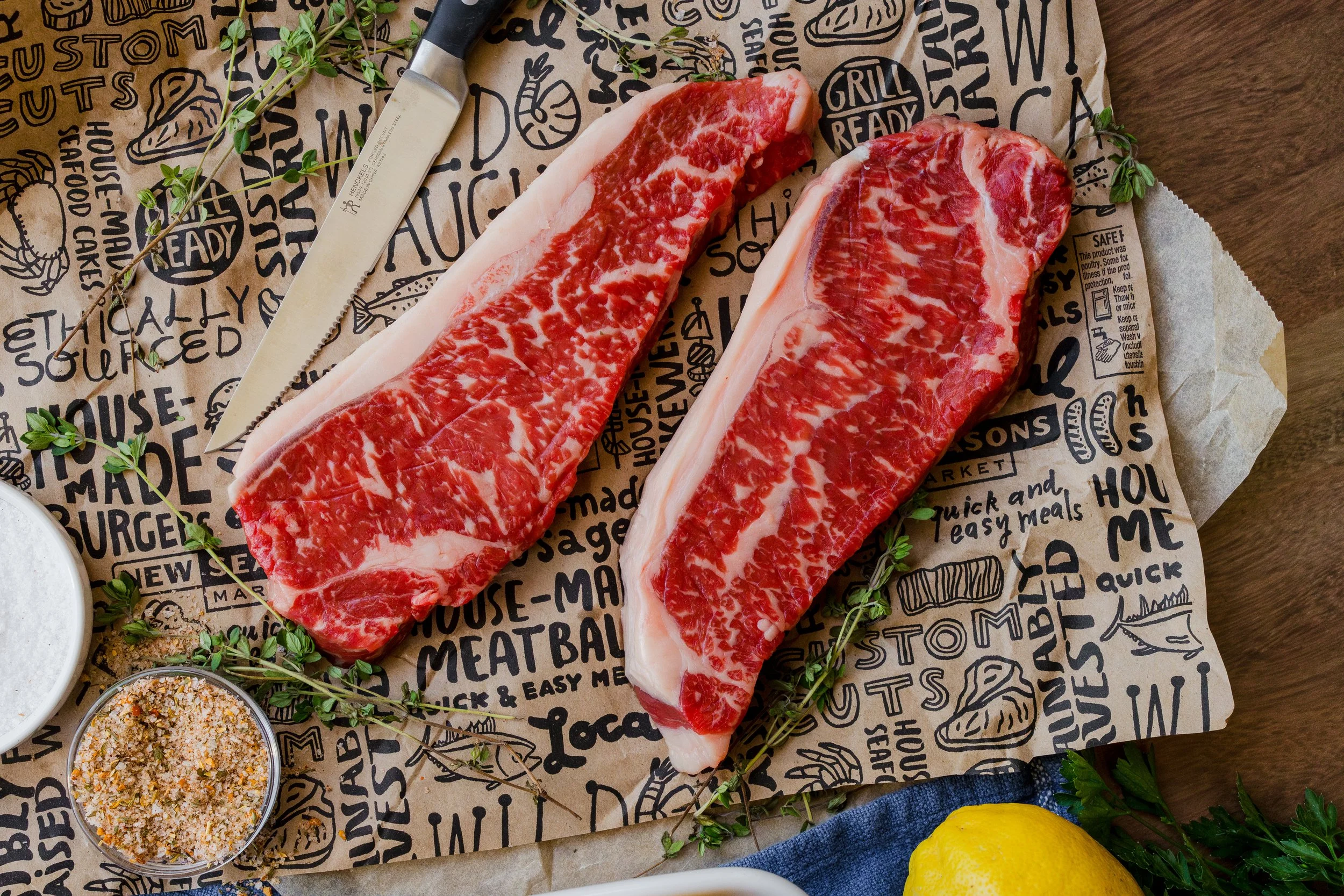 Two raw ribeye steaks with marbling on butcher paper, accompanied by a knife, sprinkled spices, herbs, and a lemon.