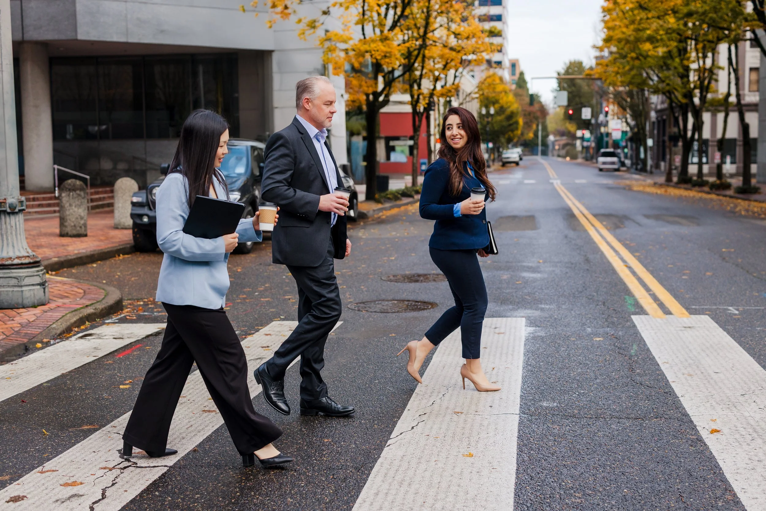 Three business professionals, two women and one man, crossing a city street on a crosswalk during autumn, holding coffee cups and documents, engaged in conversation with trees with fall foliage lining the street.