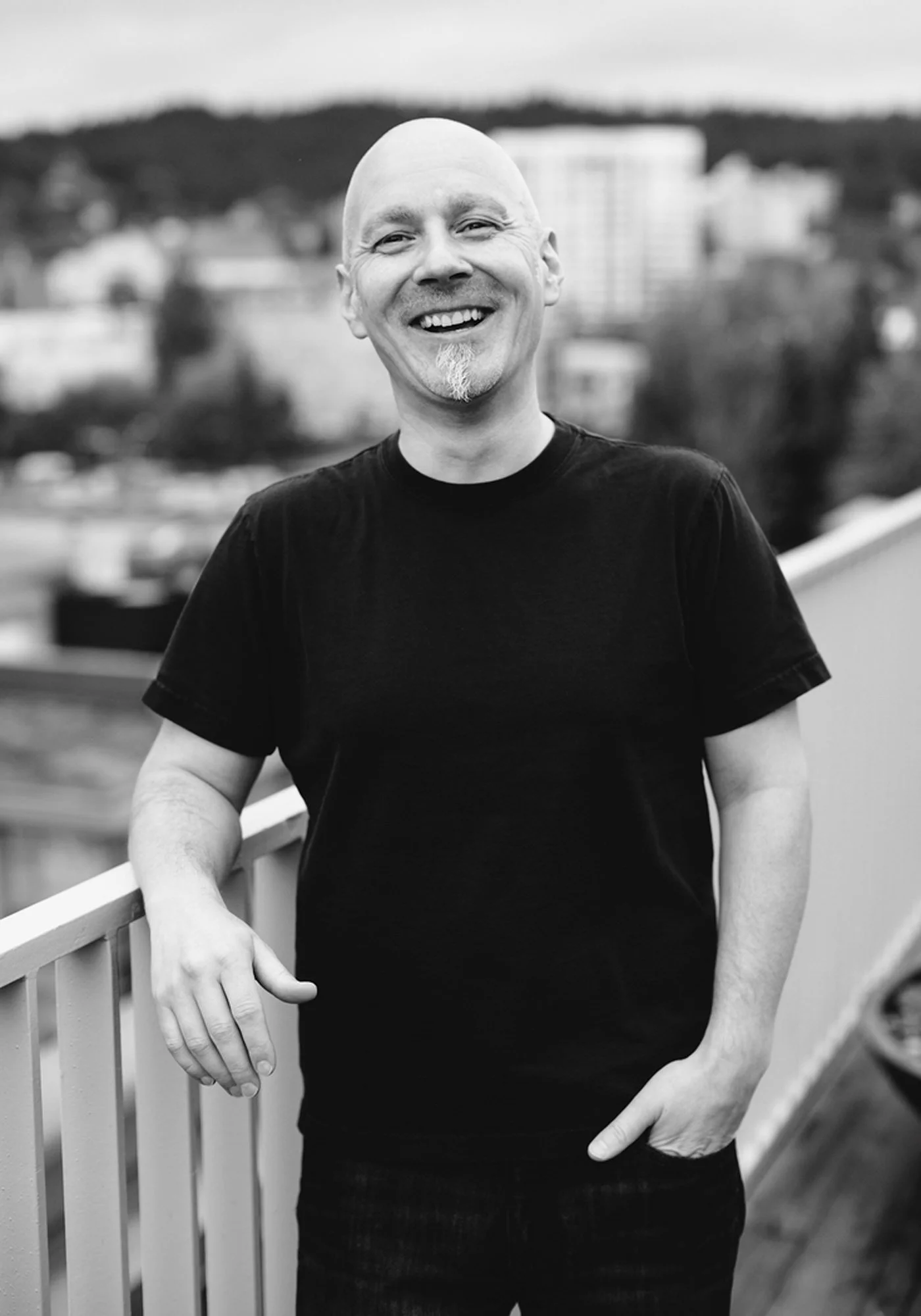 A smiling bald man with a goatee standing outdoors on a balcony with a cityscape and hills in the background in black and white.