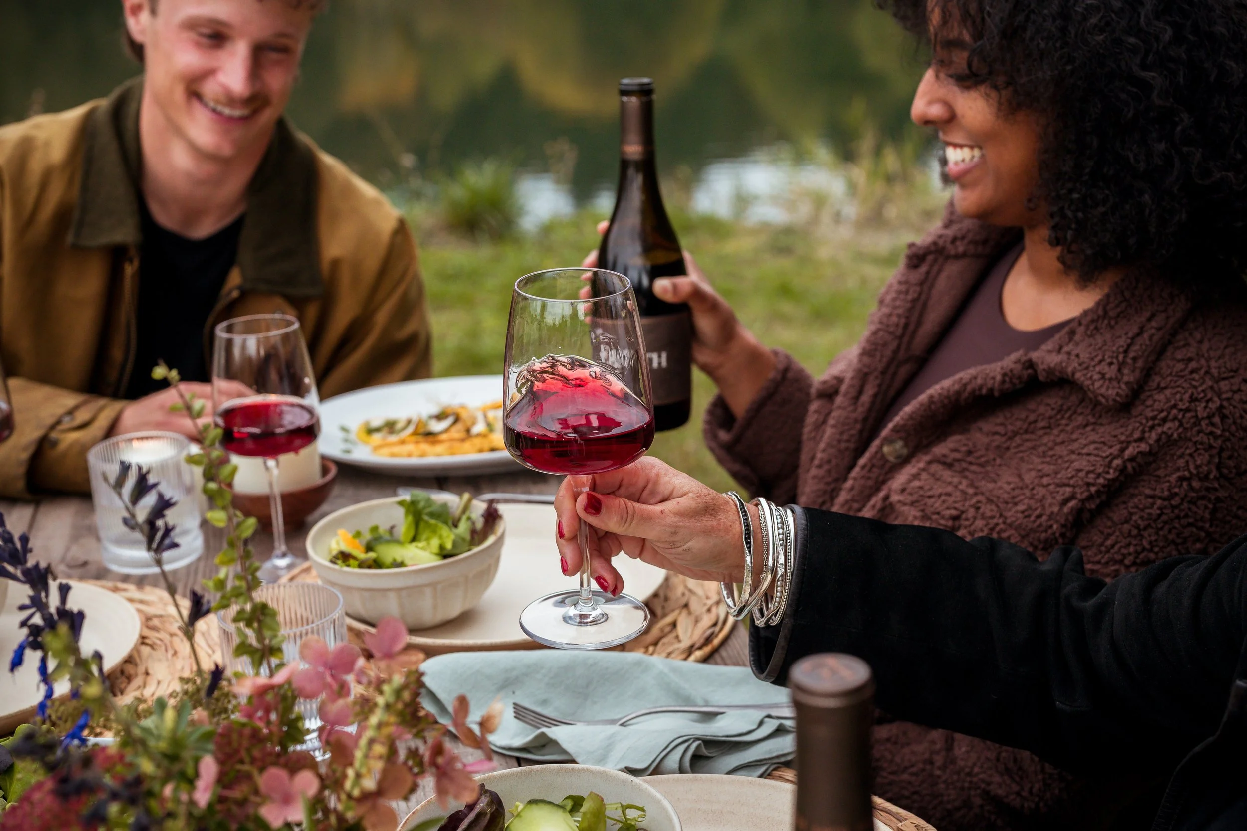 Two women and a man enjoying a meal outdoors by a lake, with one woman holding a glass of red wine and the other holding a bottle of wine. The table is set with salads, plates, and flowers.