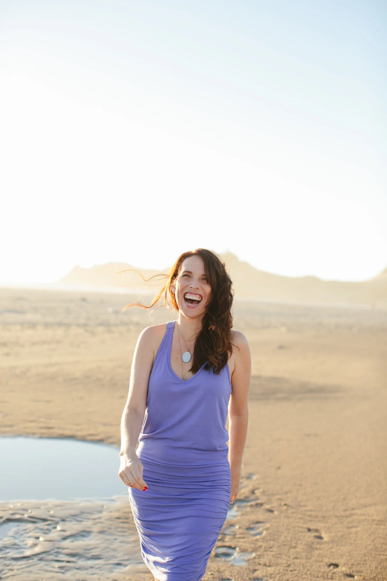 A woman in a purple dress smiling and laughing on a desert landscape with a small water pool and distant mountains under a clear sky.