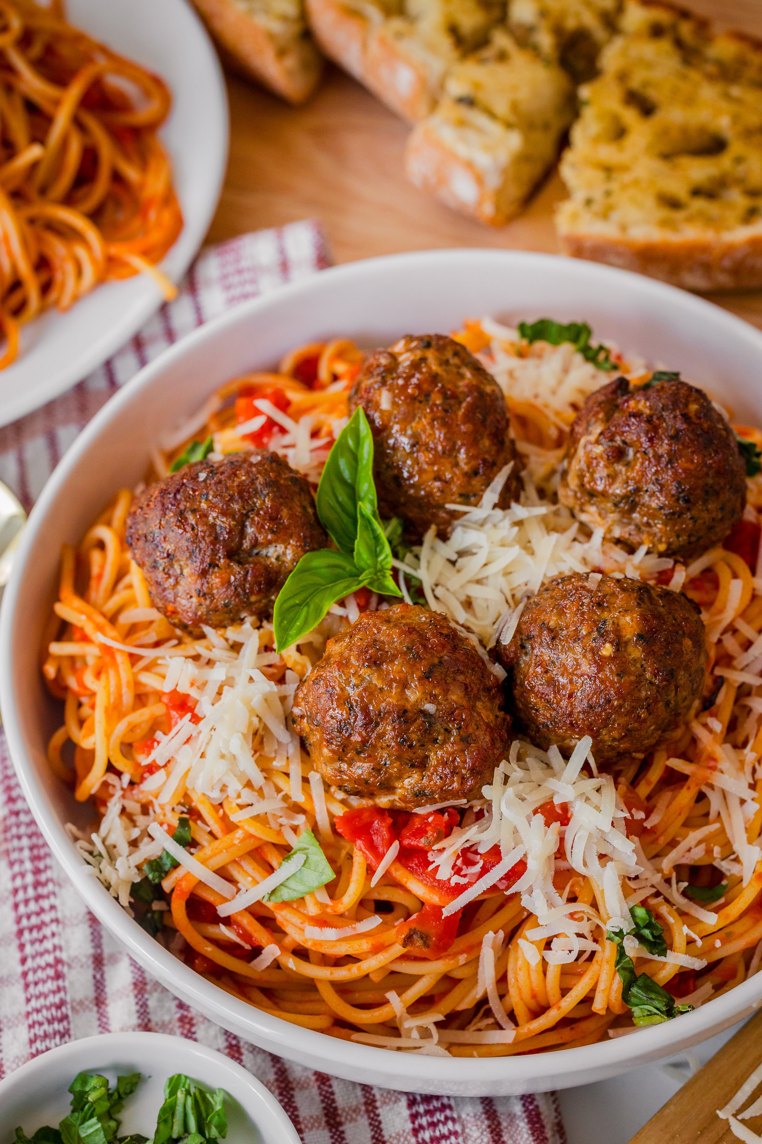 Bowl of spaghetti with meatballs topped with shredded cheese and basil, with sliced bread and a plate of additional spaghetti in the background.