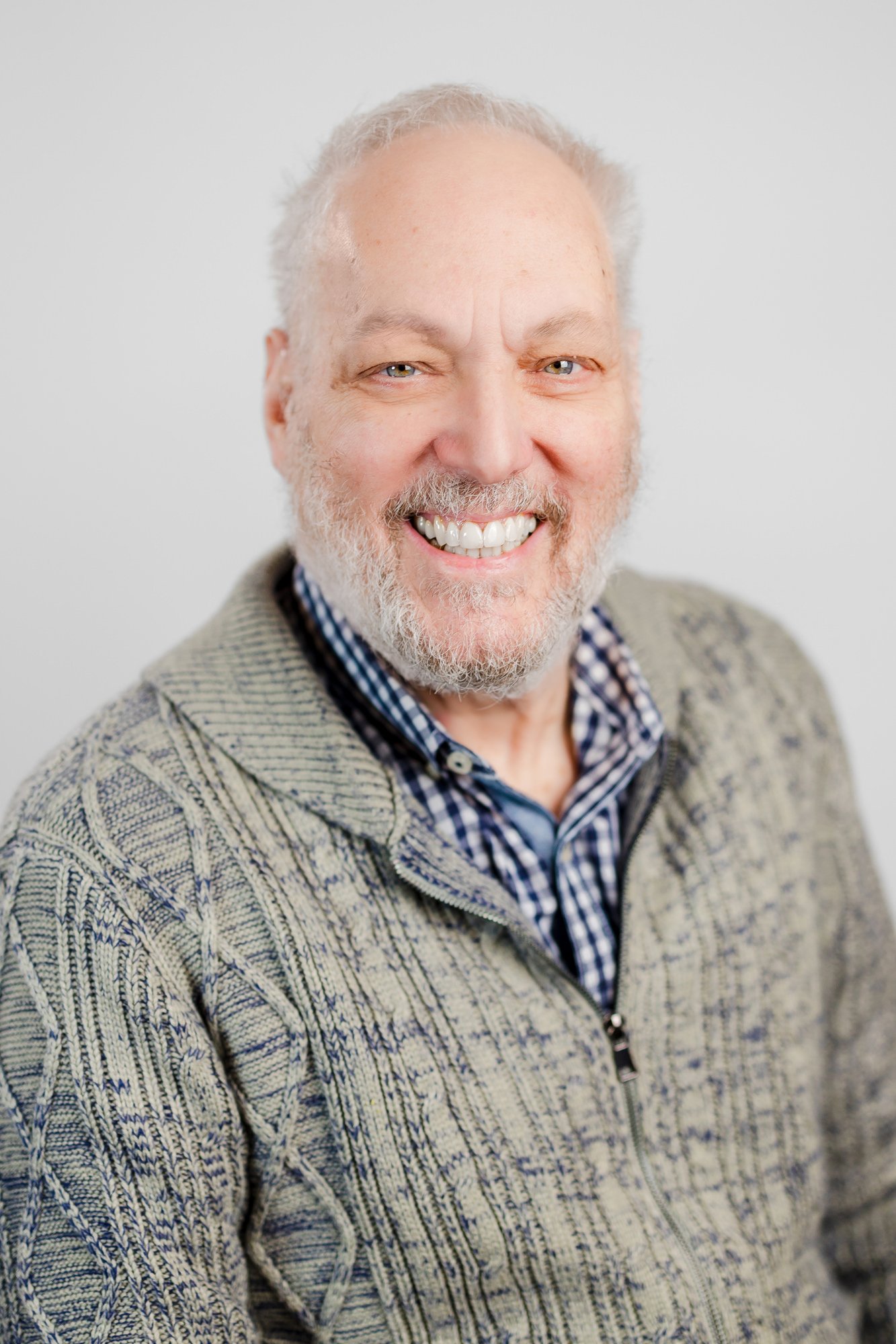 A smiling middle-aged man with a beard and light eyes, wearing a checked shirt and a textured zip-up sweater, against a plain white background.