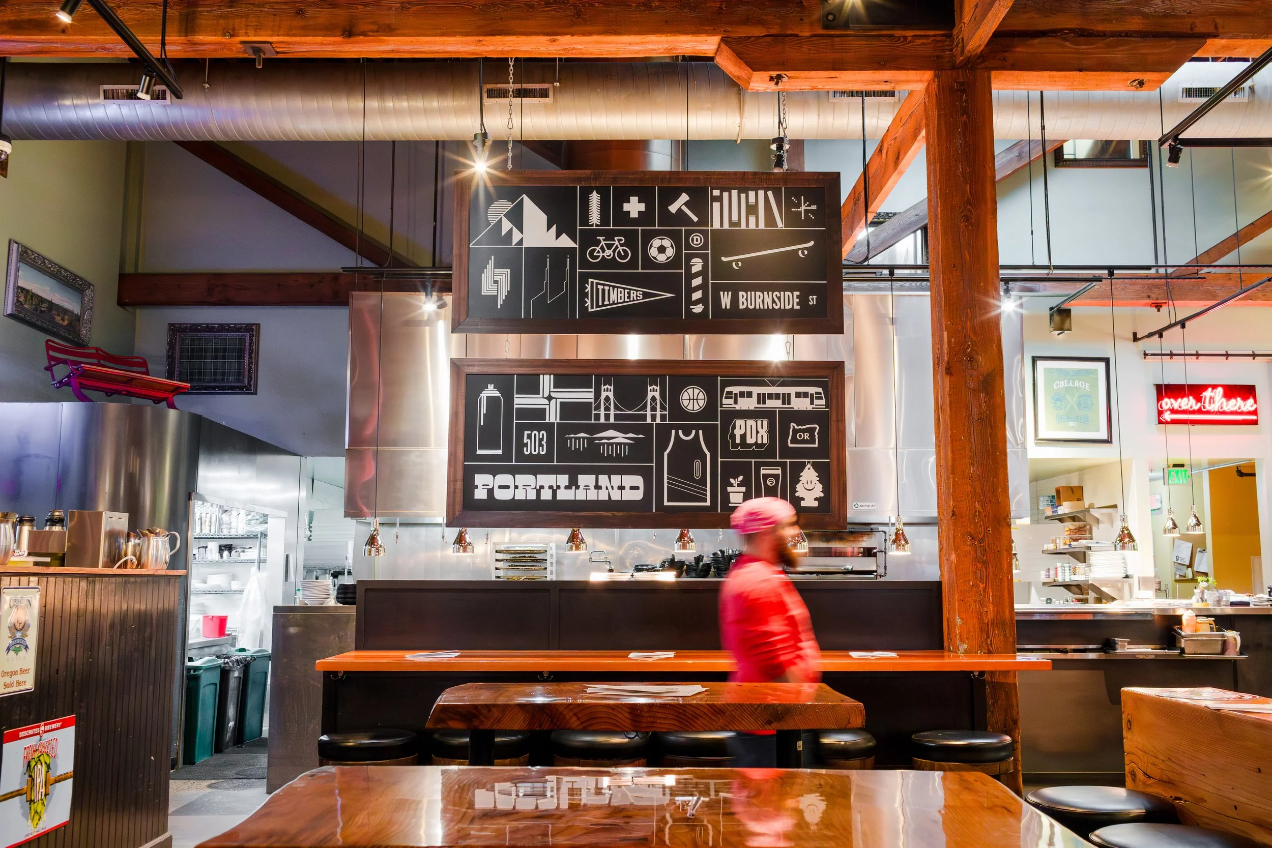 Interior of a restaurant with wooden beams and metal accents, featuring two large blackboards with white graphic symbols and text indicating Portland location and street names, with a person in pink walking past the counter.