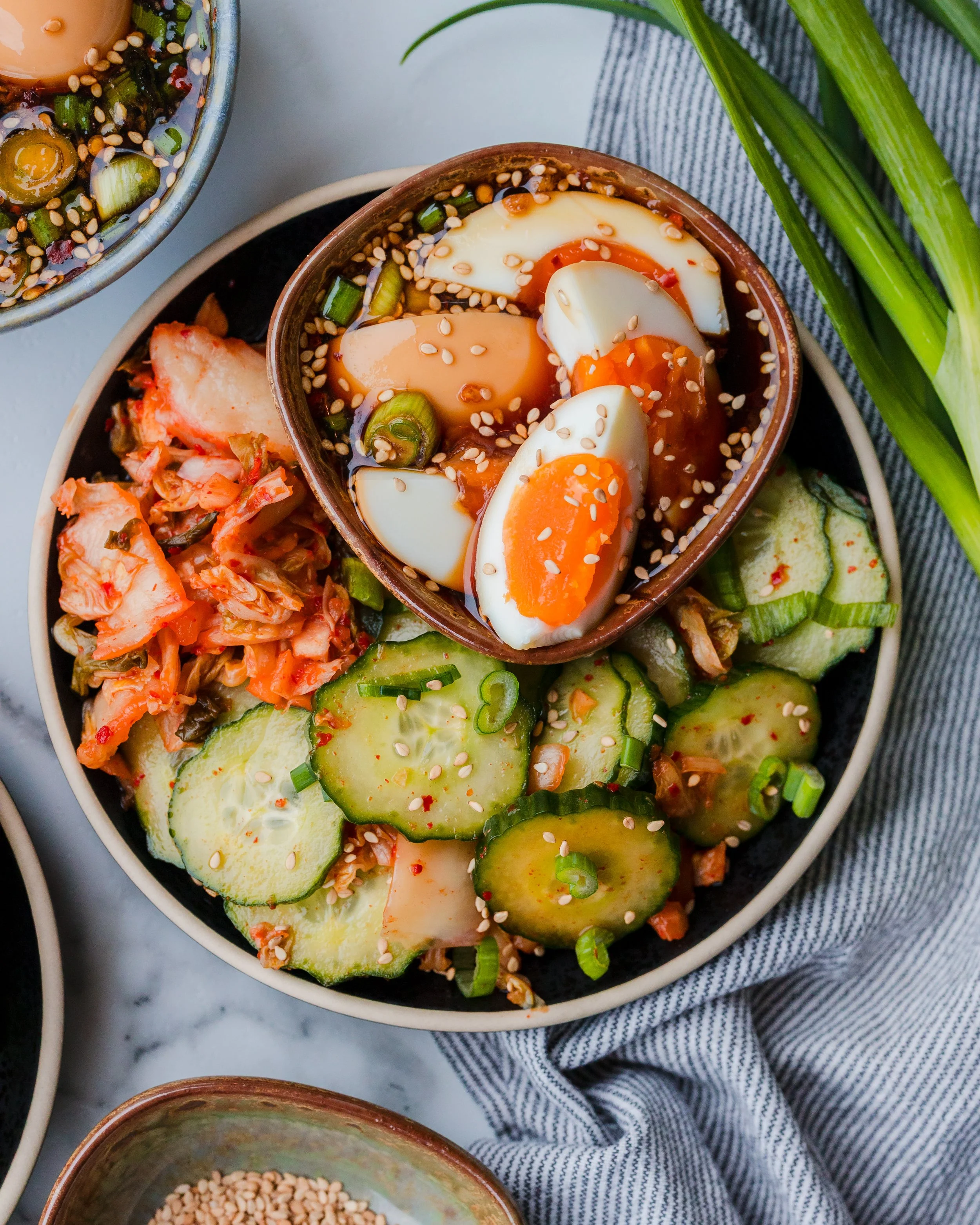 A plate of sliced cucumbers, kimchi, and a bowl of marinated boiled eggs topped with sesame seeds and scallions.
