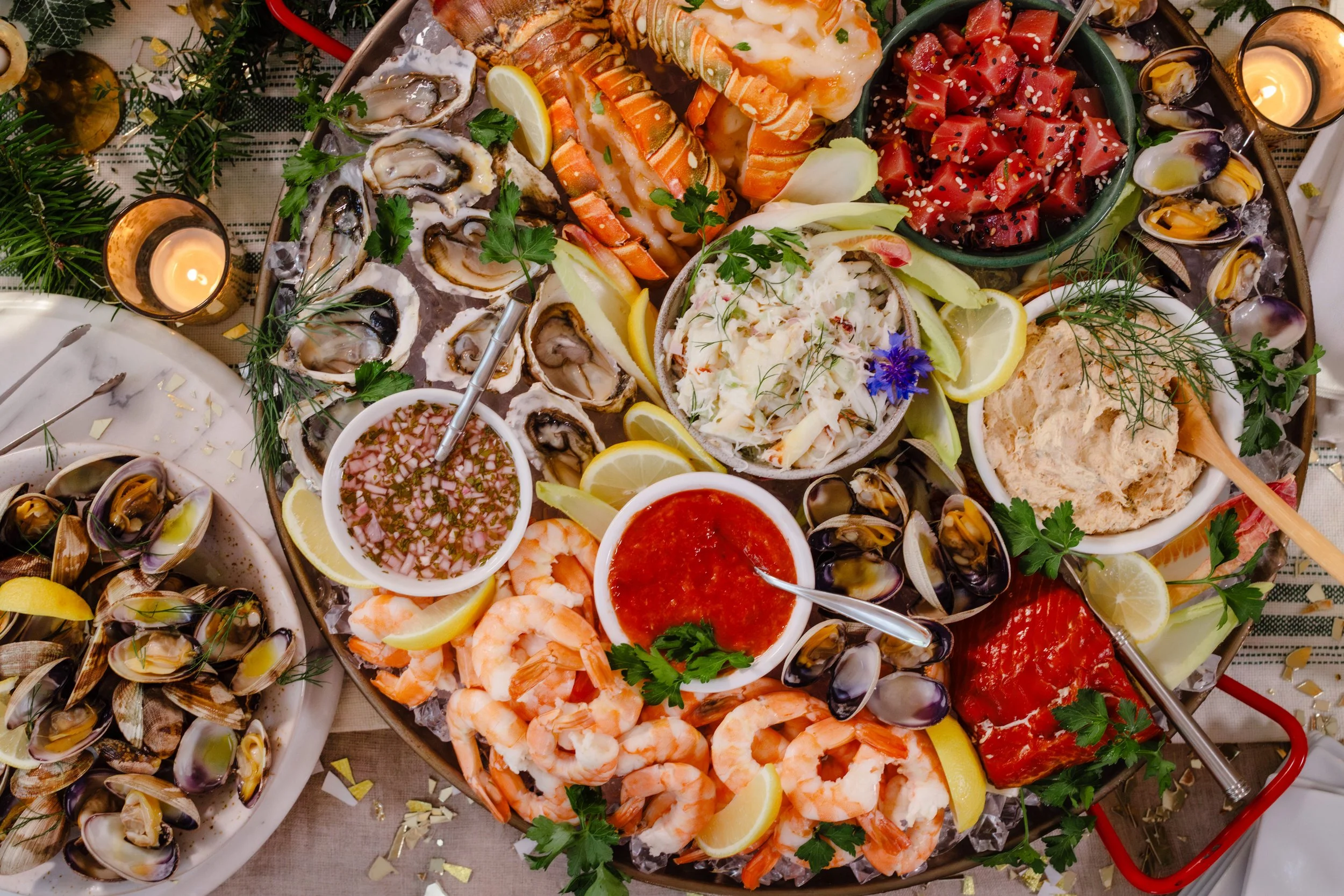 A seafood platter containing oysters on the half shell, cooked shrimp, mussels, watermelon, crab dip, coleslaw, and various dipping sauces, garnished with lemon wedges, herbs, and edible flowers.