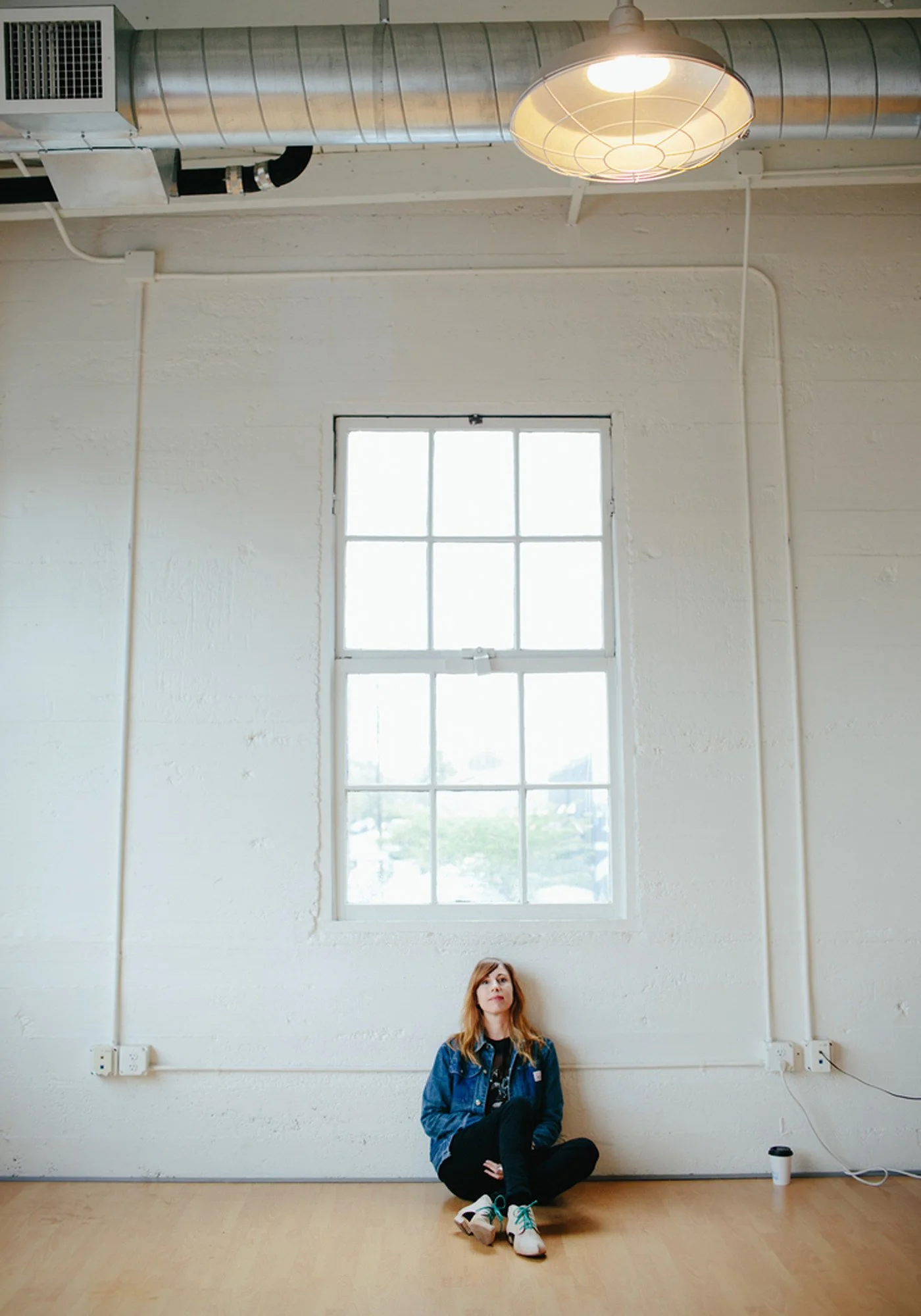 A woman with red hair, wearing a denim jacket, black pants, and white sneakers, sitting cross-legged on a wooden floor under a large window in a white-walled industrial-style room with metallic ductwork and a ceiling light.