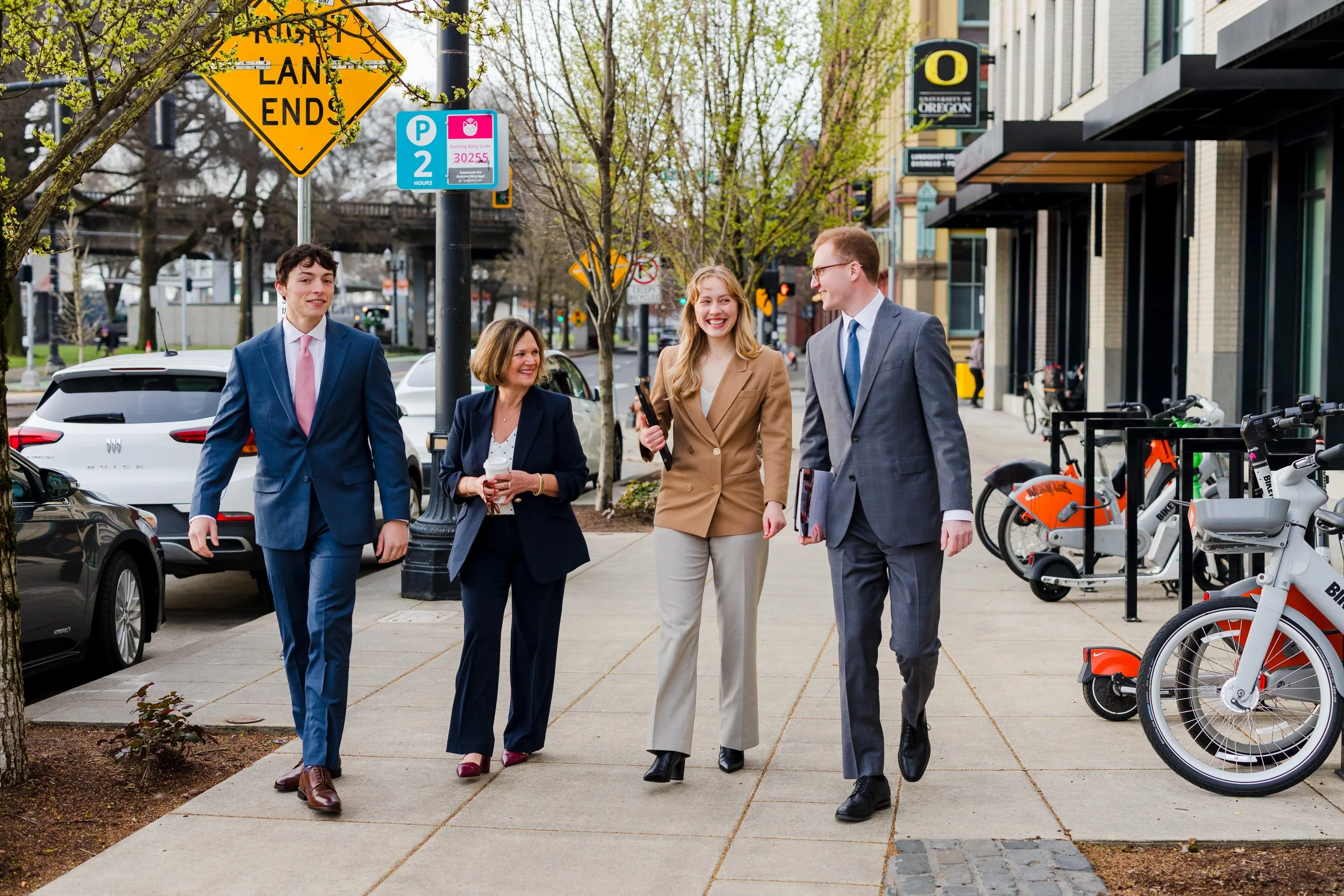 Four business professionals, two men in suits and two women, walking and talking on a city sidewalk with parked cars, bicycles, and commercial buildings in the background.