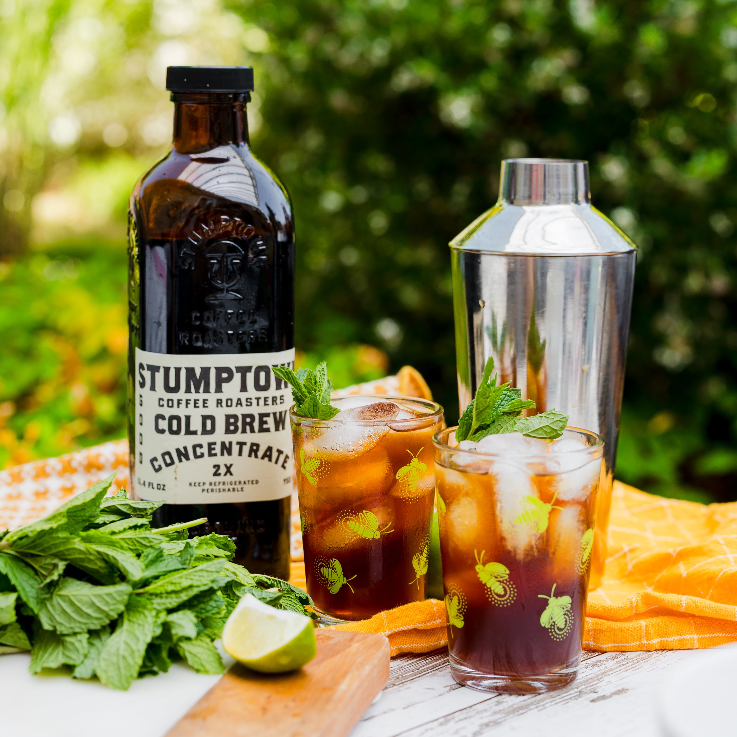 Two glasses of iced dark beverage garnished with mint leaves, a bottle of Stumptown Cold Brew Concentrate, a metal cocktail shaker, a lime wedge, mint sprigs, and a yellow tablecloth outdoors.