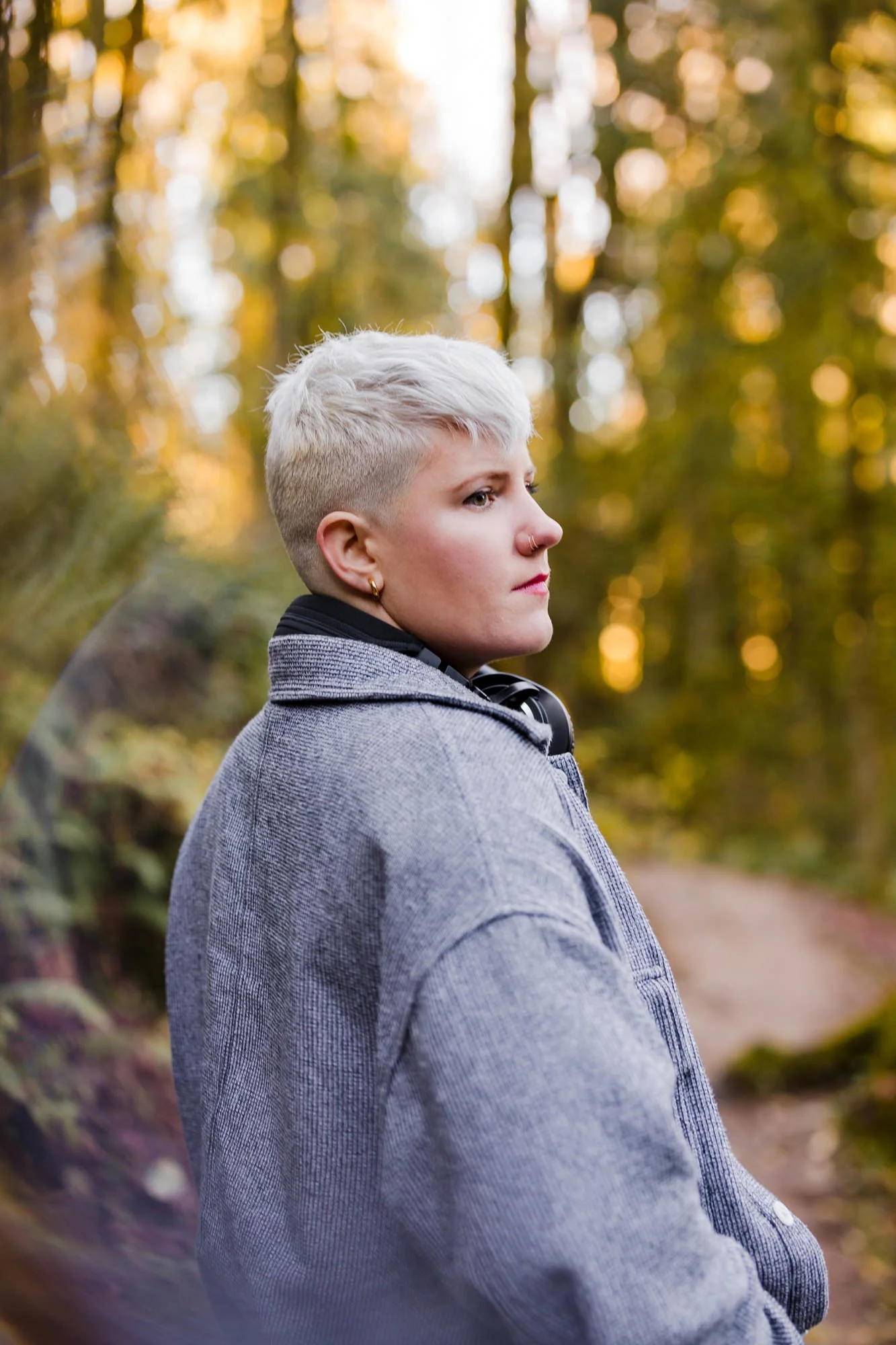 A person with short, platinum blonde hair, wearing a gray coat, standing outdoors in a forested area during autumn, with blurred foliage and warm sunlight in the background.