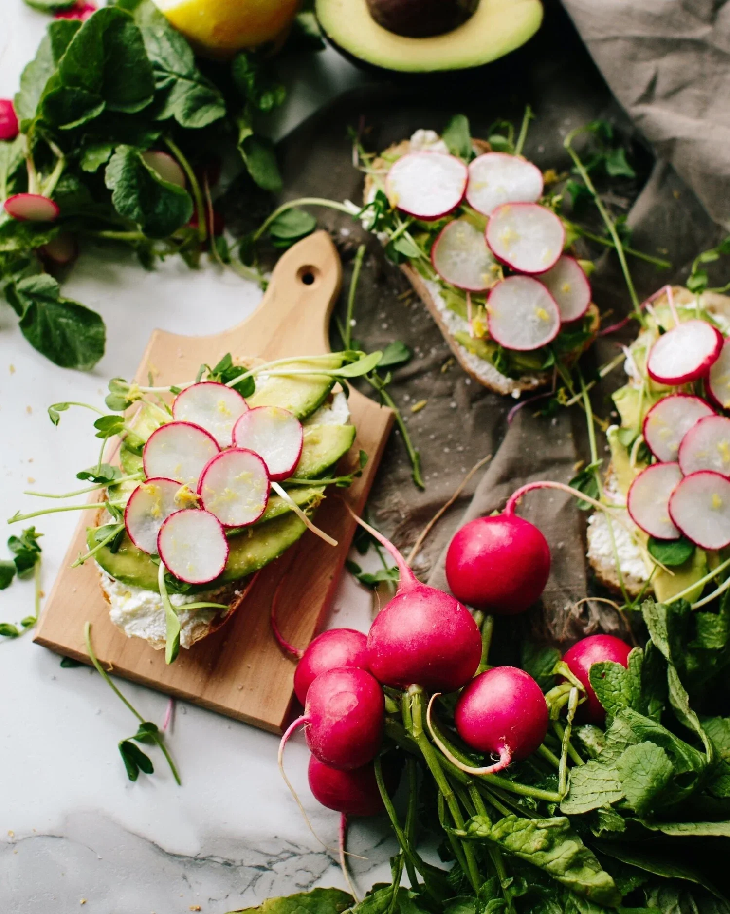 Radish-topped open-faced sandwiches on a cutting board and marble surface, with radishes, avocado, and greens nearby.