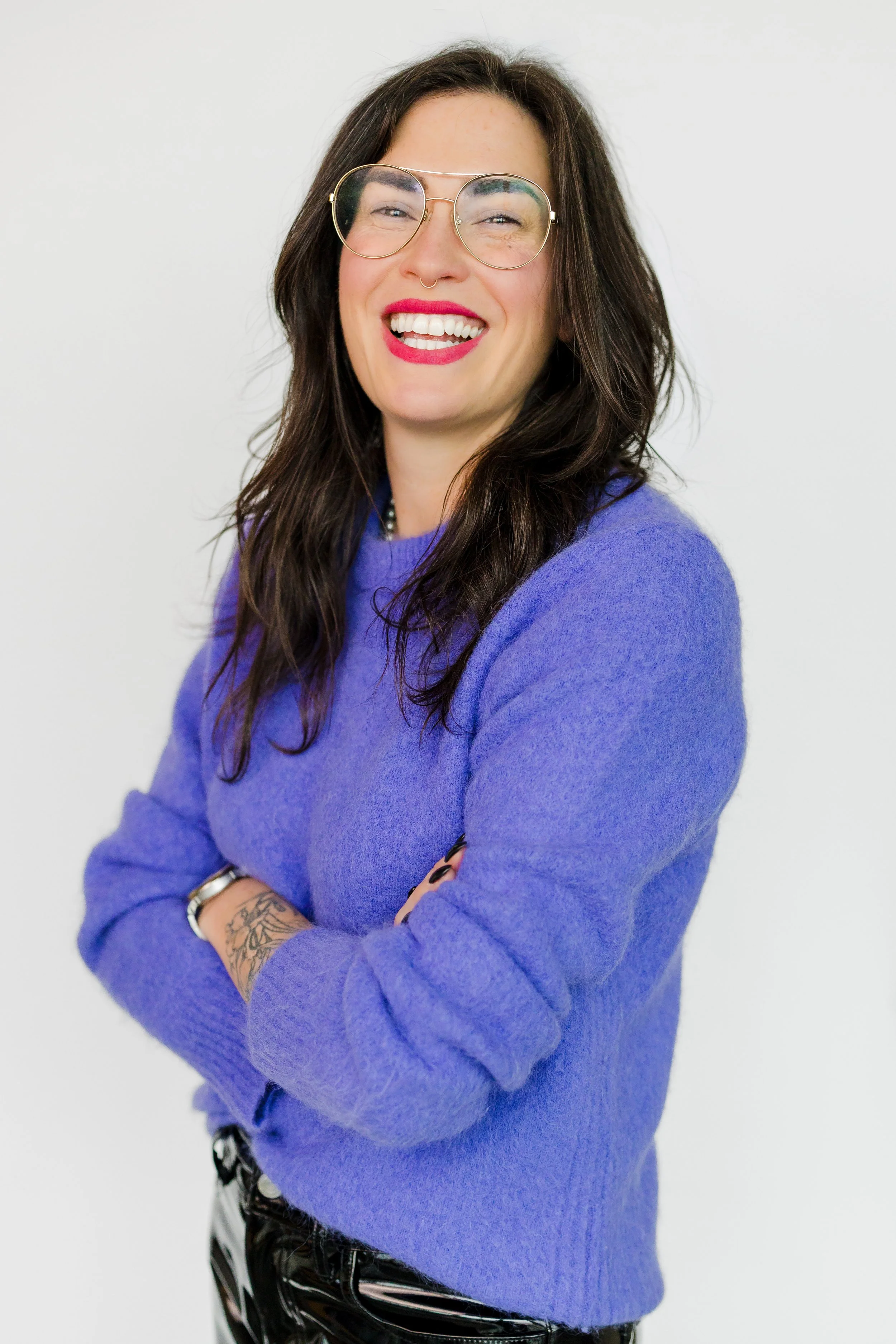 A woman with long dark hair, wearing glasses and a purple sweater, smiling with arms crossed against a plain white background.