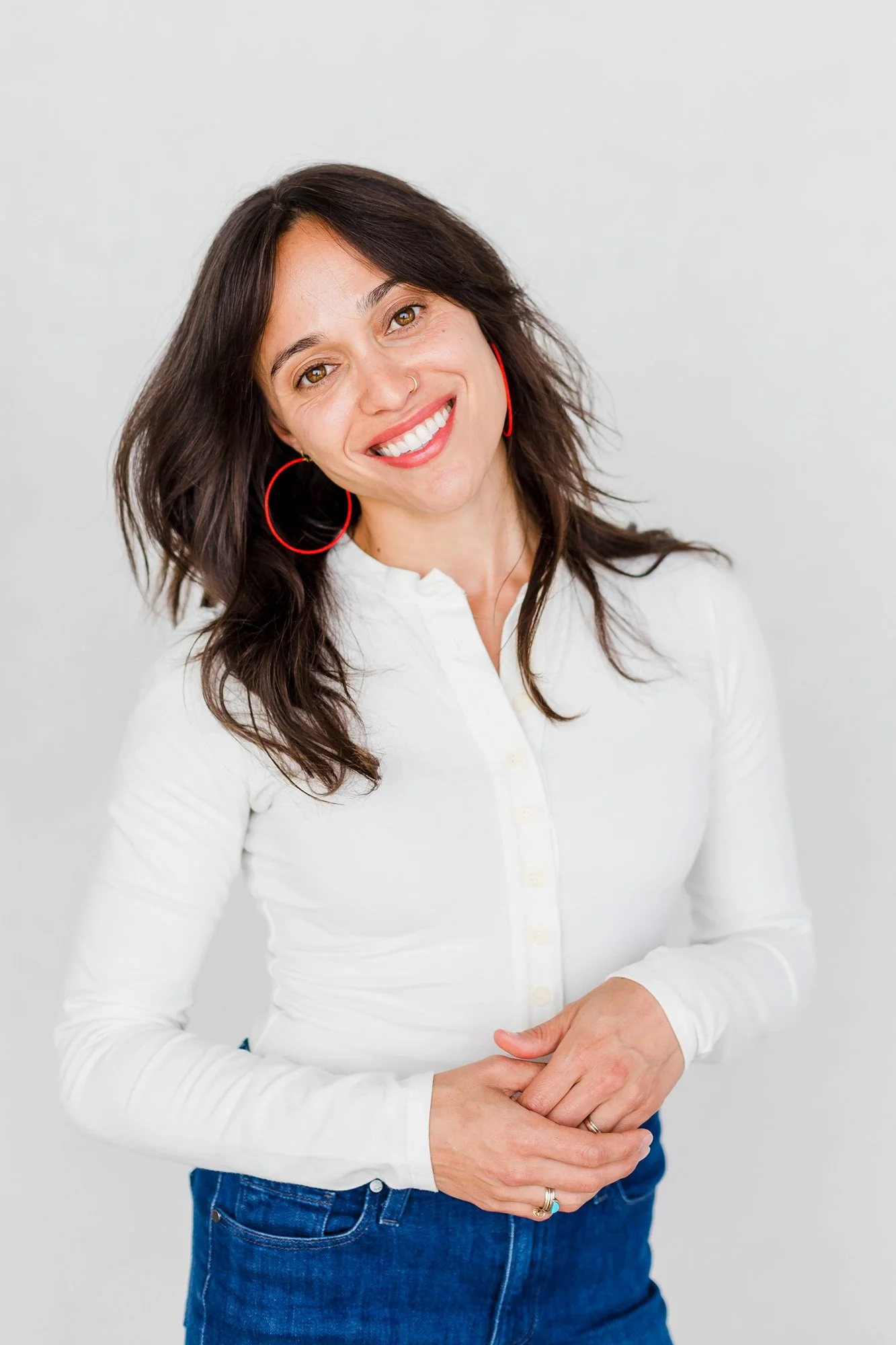 A woman with long brown hair, wearing a white button-up shirt, blue jeans, and red hoop earrings, standing against a plain white background, smiling at the camera.