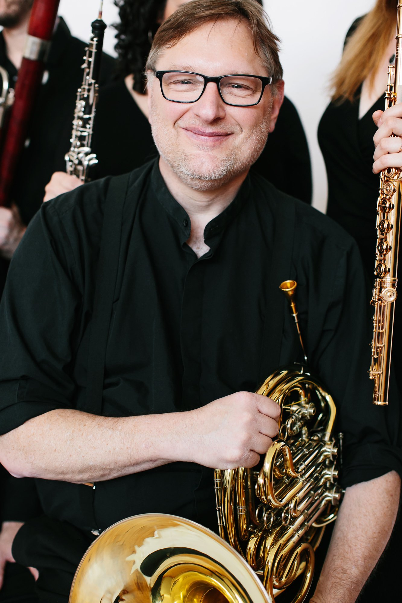 A smiling man with glasses and a beard holding a brass saxophone in front of a group of musicians.