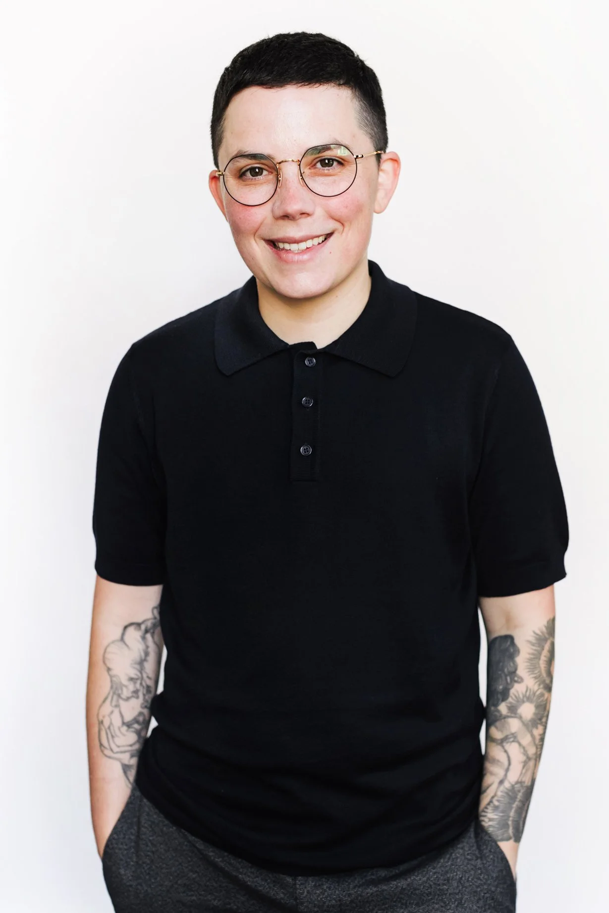 Portrait of a person with short dark hair, wearing glasses, a black polo shirt, smiling, standing against a plain white background.