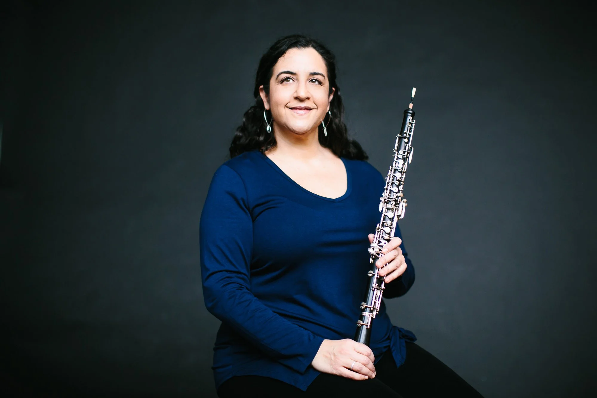 A woman with dark curly hair wearing a blue long-sleeve shirt, holding a clarinet, sitting against a dark background.