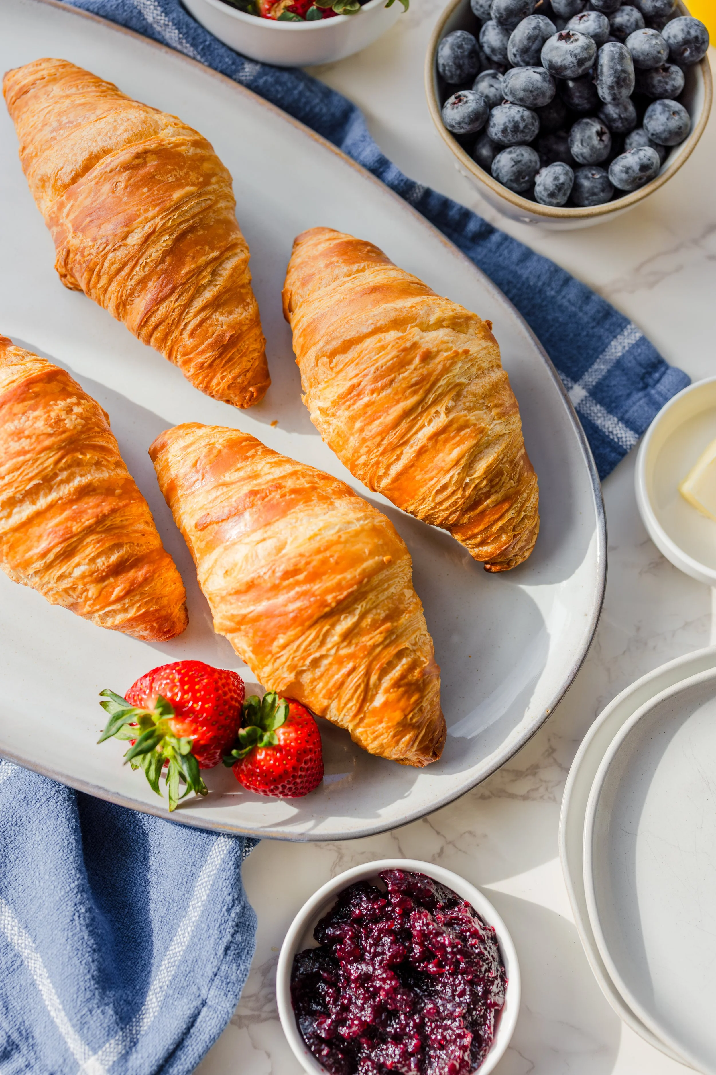 Four golden-brown croissants on a white serving platter with fresh strawberries at the corner, surrounded by bowls of blueberries, berry jam, butter, and a white dish with empty space, set on a marble surface with a blue cloth napkin nearby.