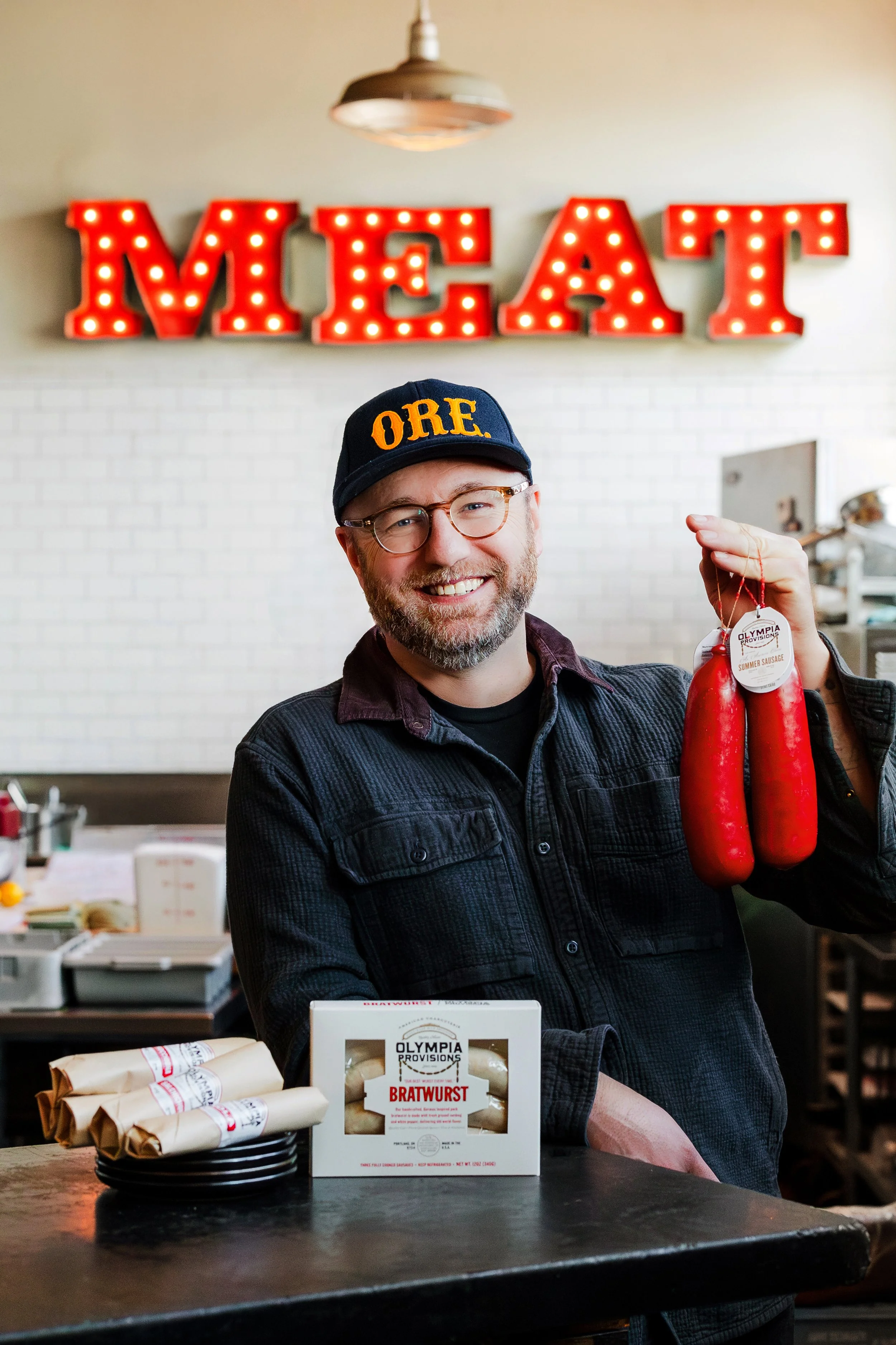 A man with glasses and a beard holding two red sausages with a tag, standing in front of a table with wrapped sausages and a box of brats, in a room with a white brick wall and red illuminated letters spelling 'MEAT' in the background.