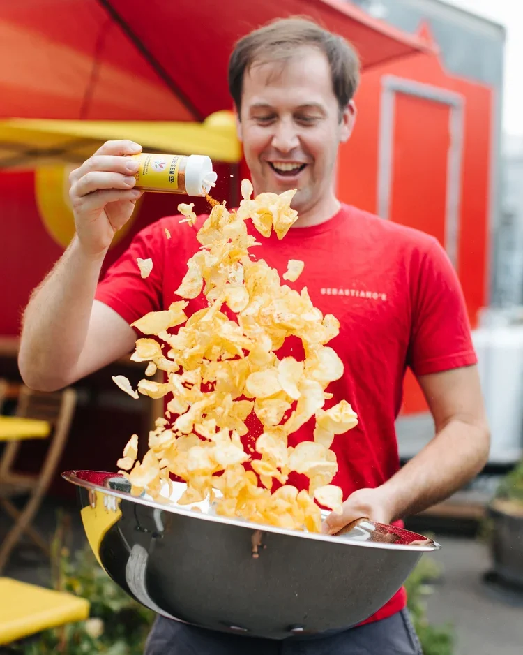 A man smiling and pouring seasoning from a yellow container onto a large bowl of potato chips in an outdoor setting with a red food truck and yellow chairs in the background.