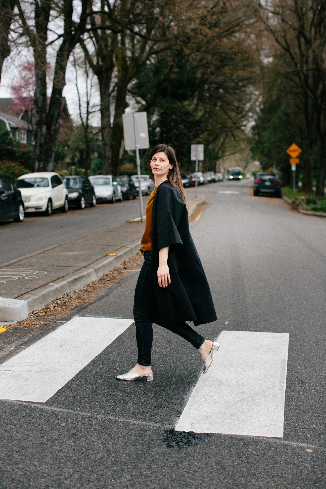 A woman crossing a street at a pedestrian crosswalk in a residential neighborhood, with parked cars and trees in the background.
