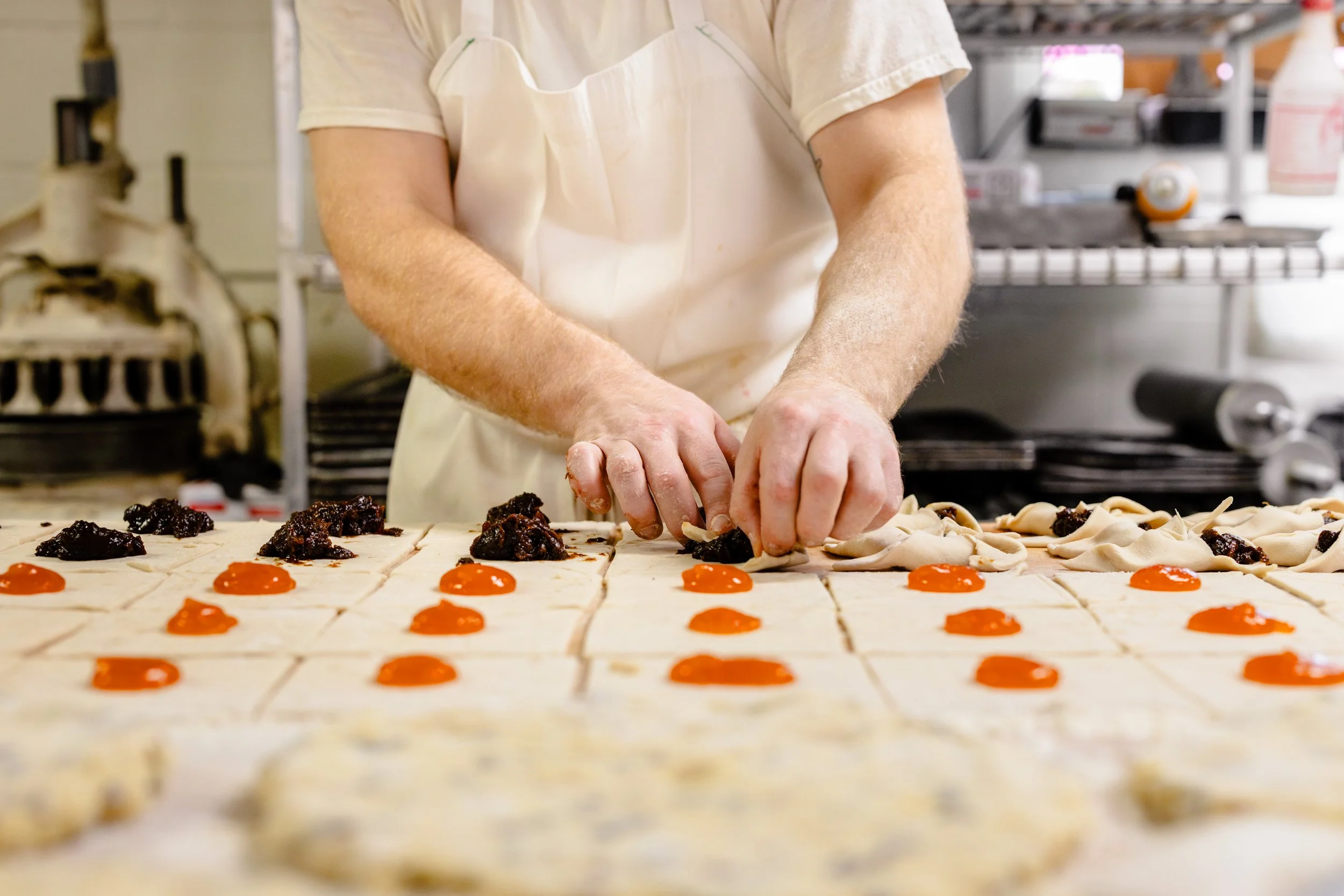 Someone preparing pastries with dollops of orange jam and dark fruit filling on a baking tray in a kitchen.
