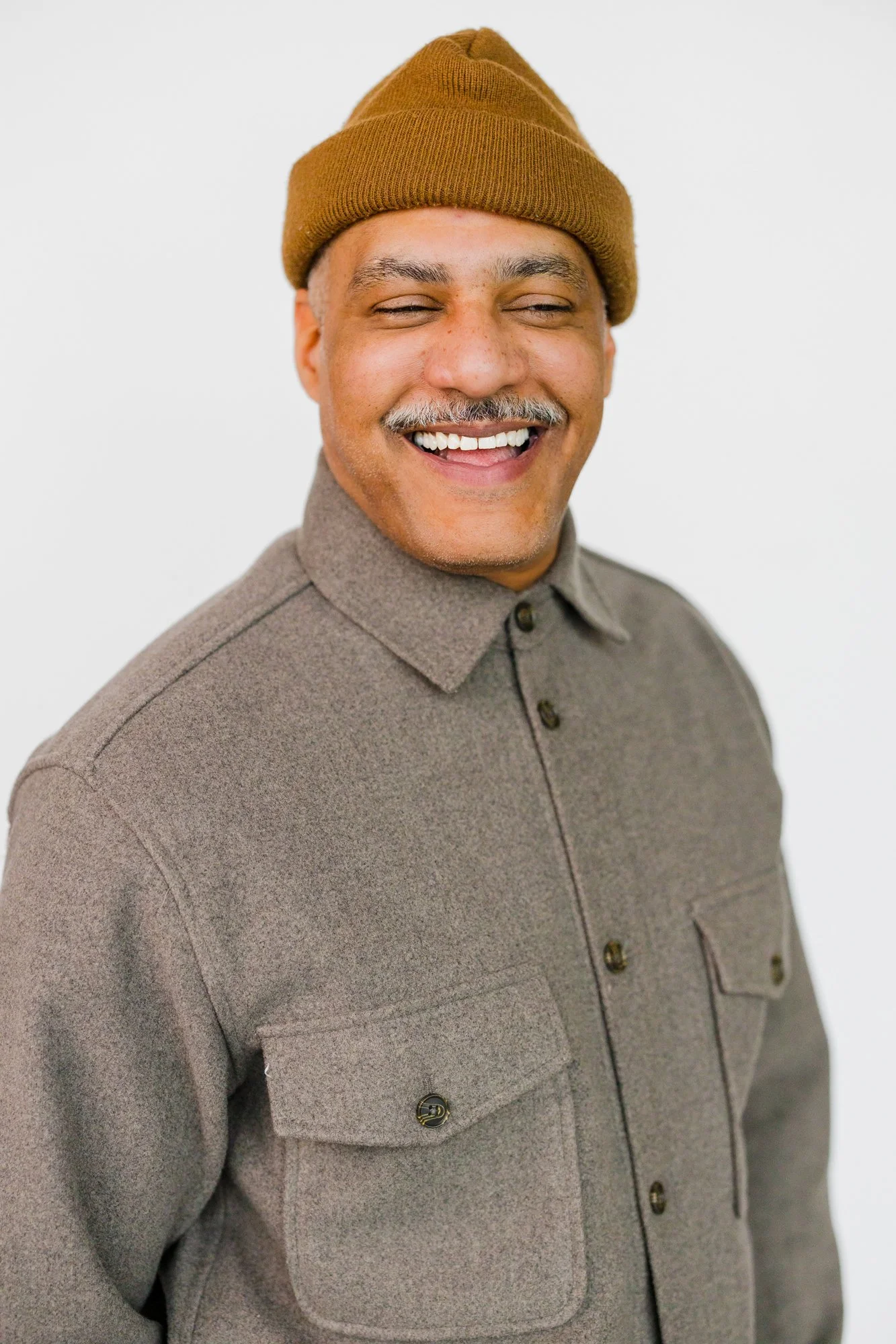 A smiling man wearing a brown beanie, a gray button-up shirt, and standing against a white background.