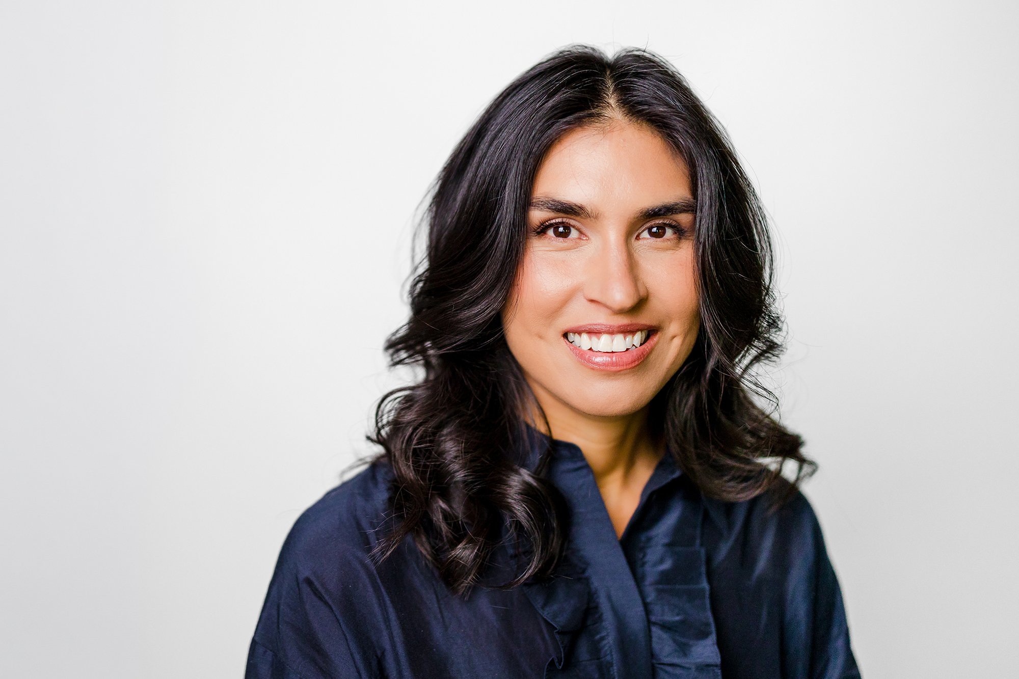 A professional woman with long, dark, curly hair smiling at the camera, wearing a dark blue shirt.