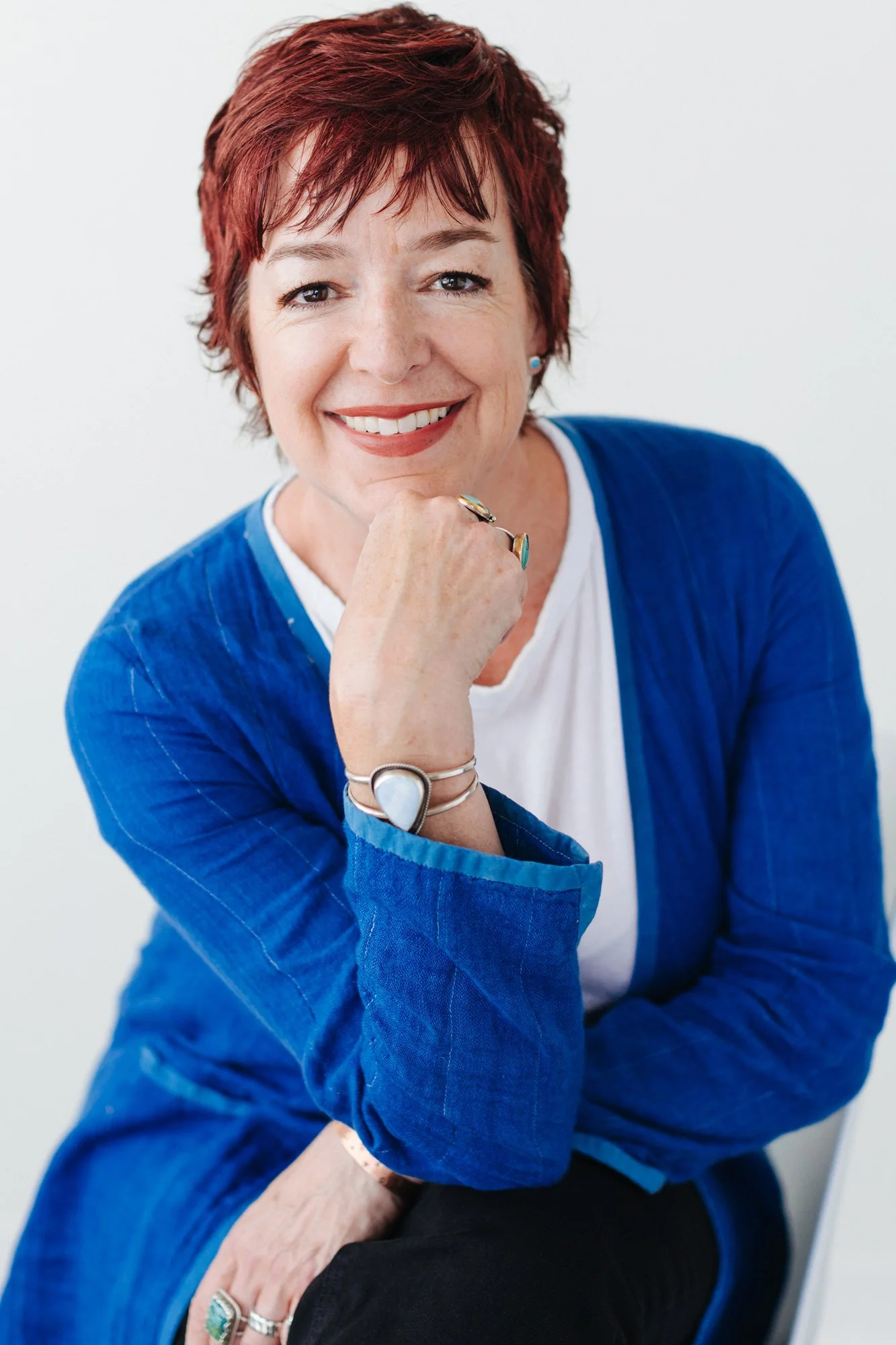 A woman with short red hair smiling, wearing a blue jacket over a white top, sitting with her hand resting on her chin, wearing multiple rings and bracelets, against a plain light background.