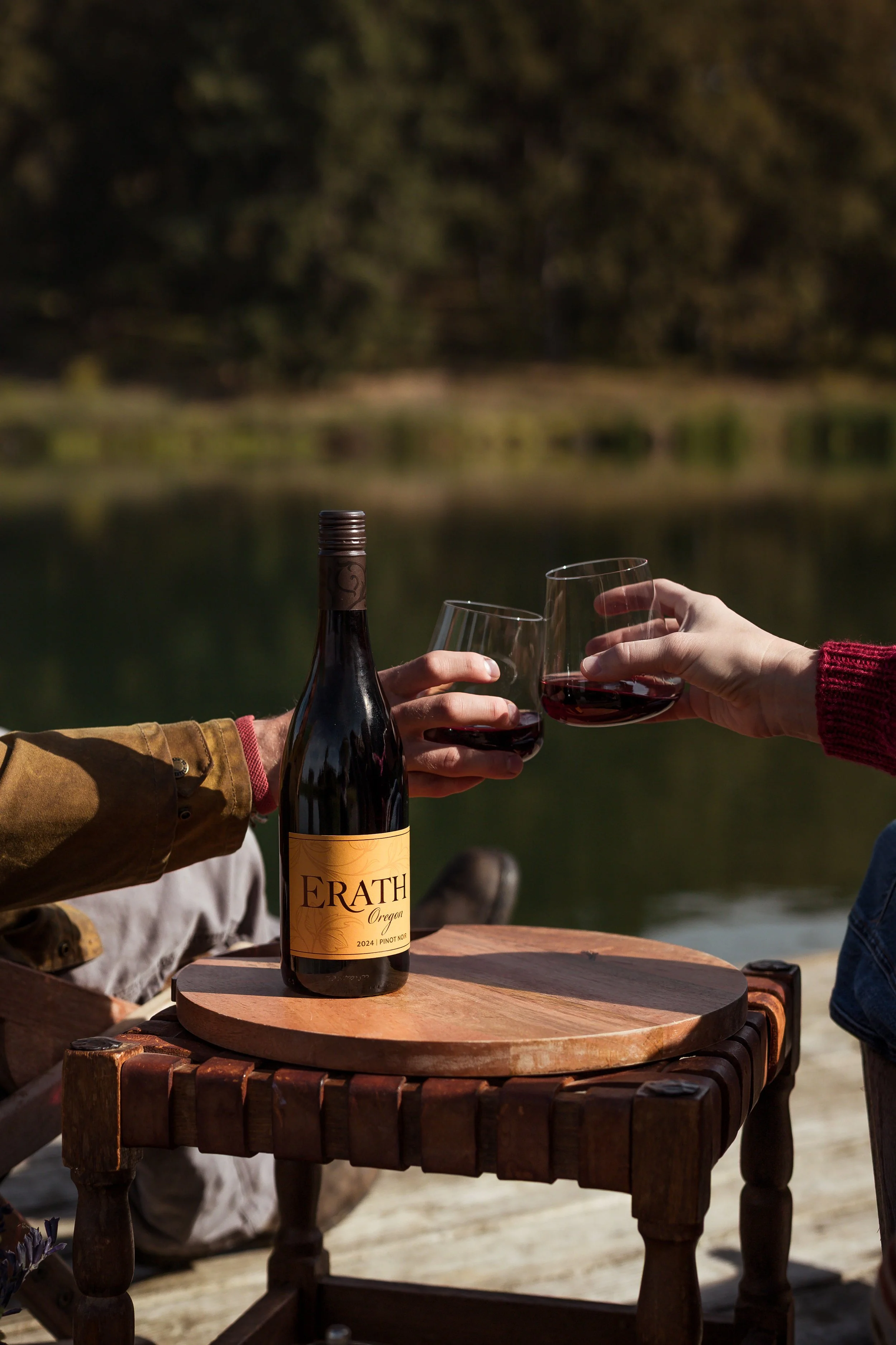 Two people clinking glasses of red wine outdoors, with a bottle of Erath Pinot Noir on a wooden table near a body of water and trees in the background.