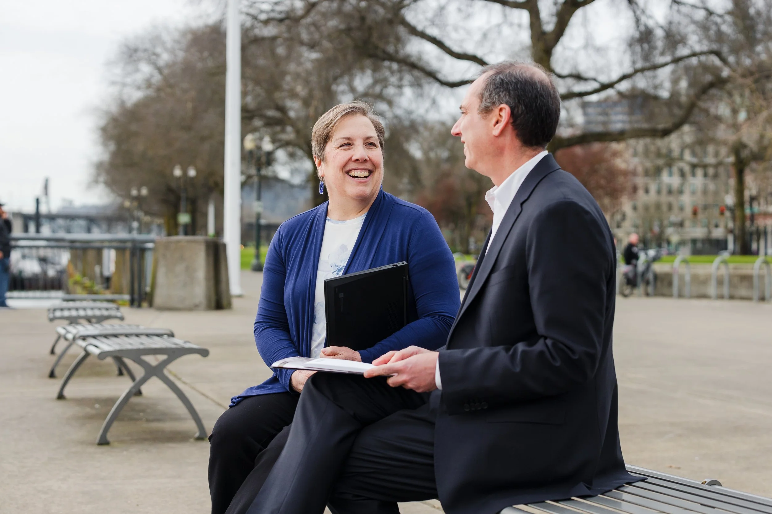 A woman and a man are sitting on a park bench, engaged in a conversation, smiling at each other. The woman is holding a black folder and papers, and the man is holding a notebook. They are dressed in business attire. In the background, there are tree
