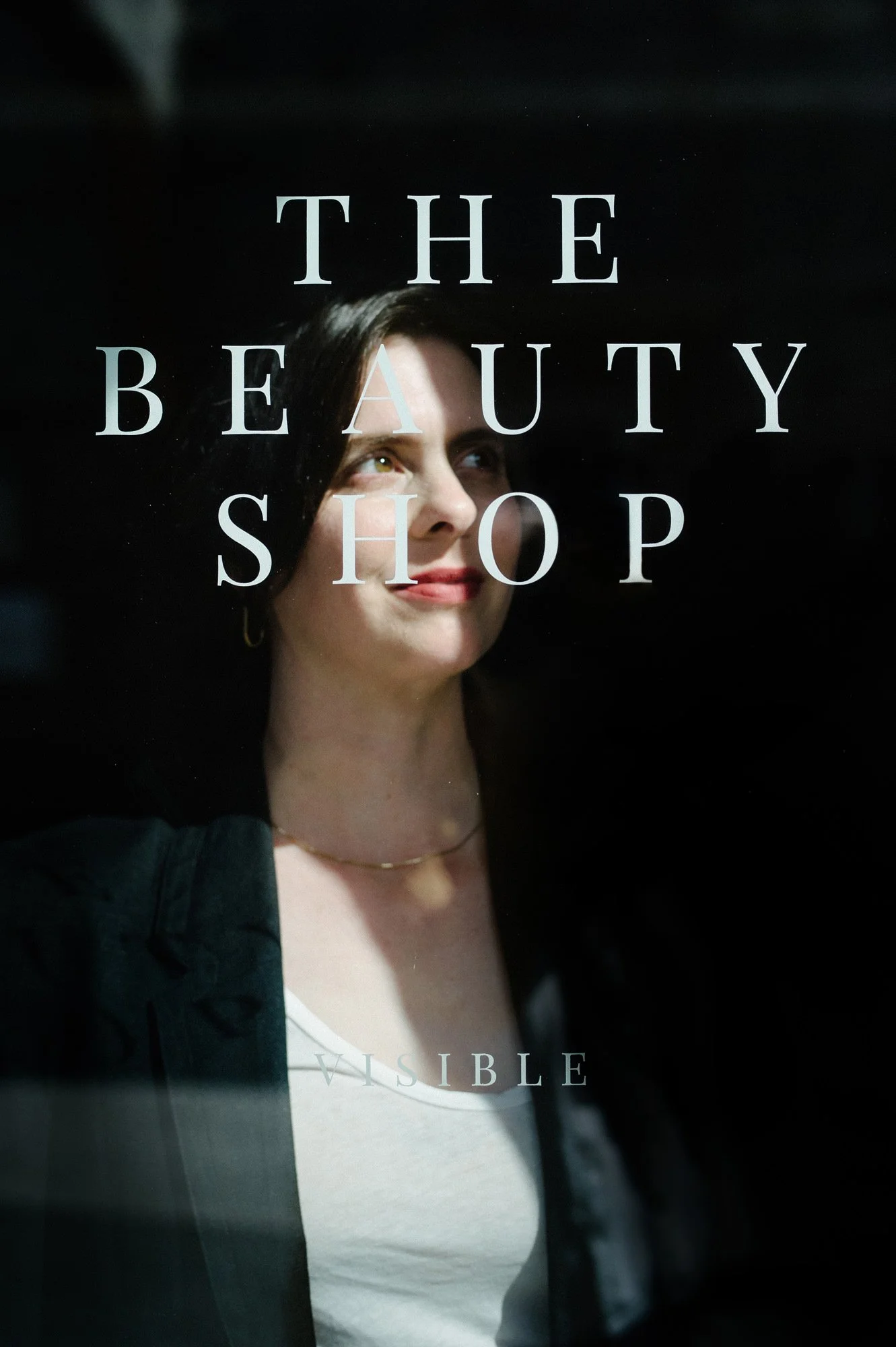 A woman with short dark hair looking to her right, behind a glass window with the words 'The Beauty Shop' and 'Visible' written on it.