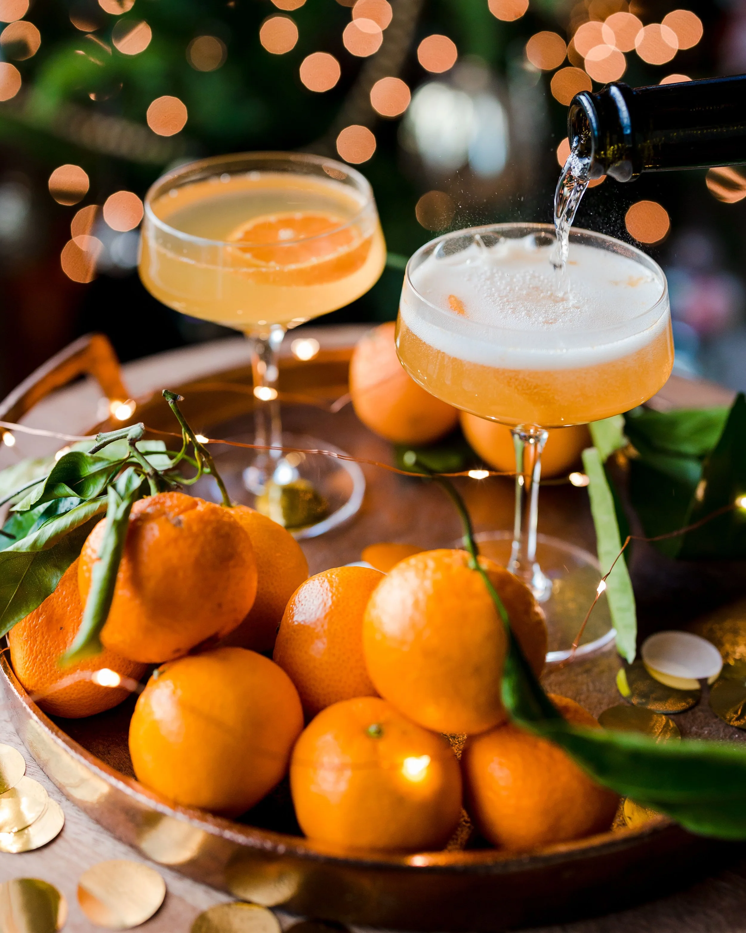 Two champagne glasses with orange slices, one being filled with sparkling wine, surrounded by oranges and green leaves on a wooden tray, with a blurred background of warm lights.