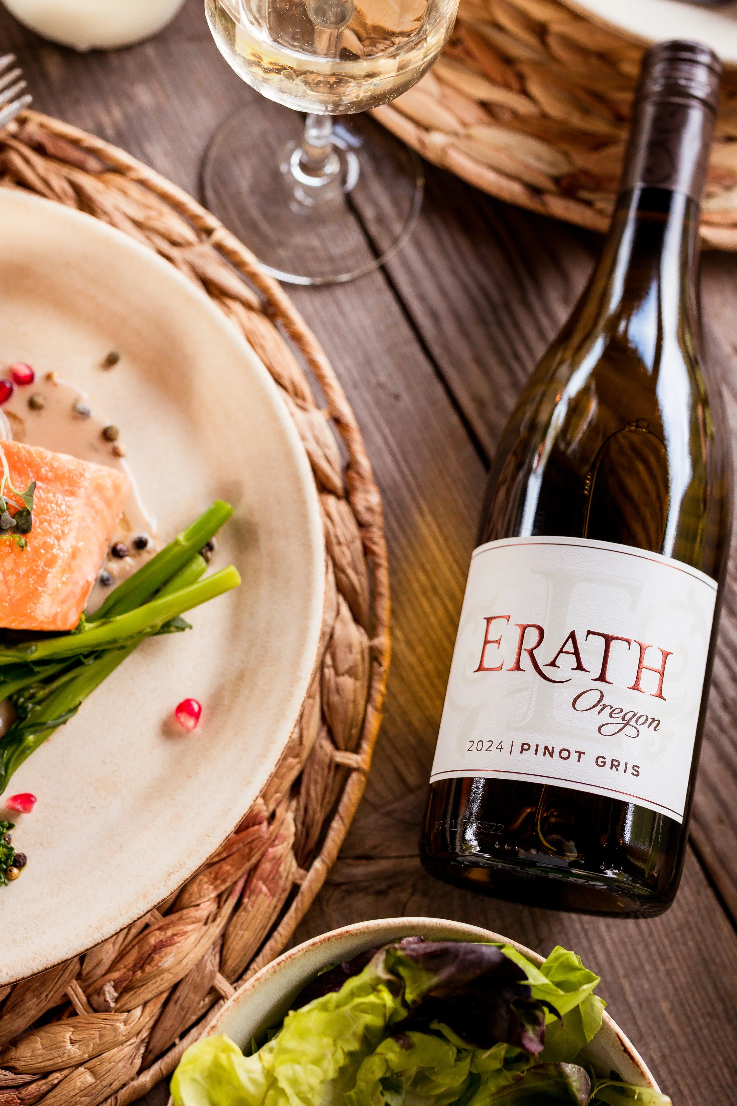 A bottle of Erath Oregon Pinot Gris wine, a glass of white wine, a plate with salmon, green beans, and peppercorns, and a bowl of mixed greens on a wooden table.