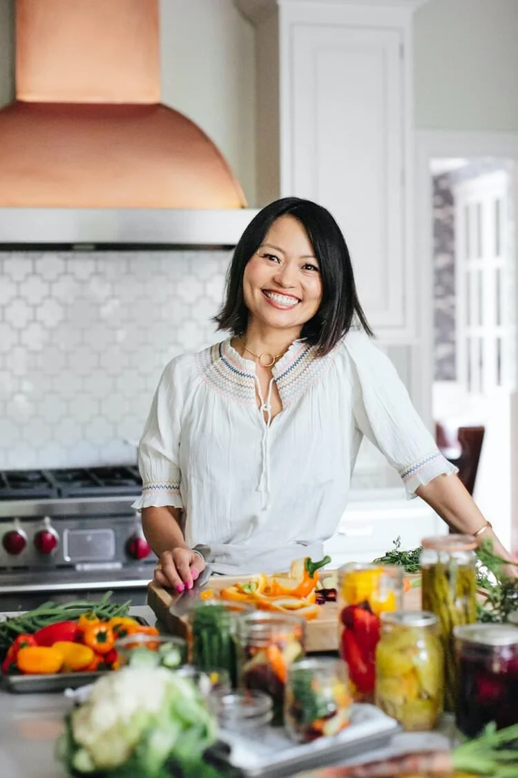 Woman preparing vegetables in a bright kitchen with a variety of fresh produce and jars on the countertop.