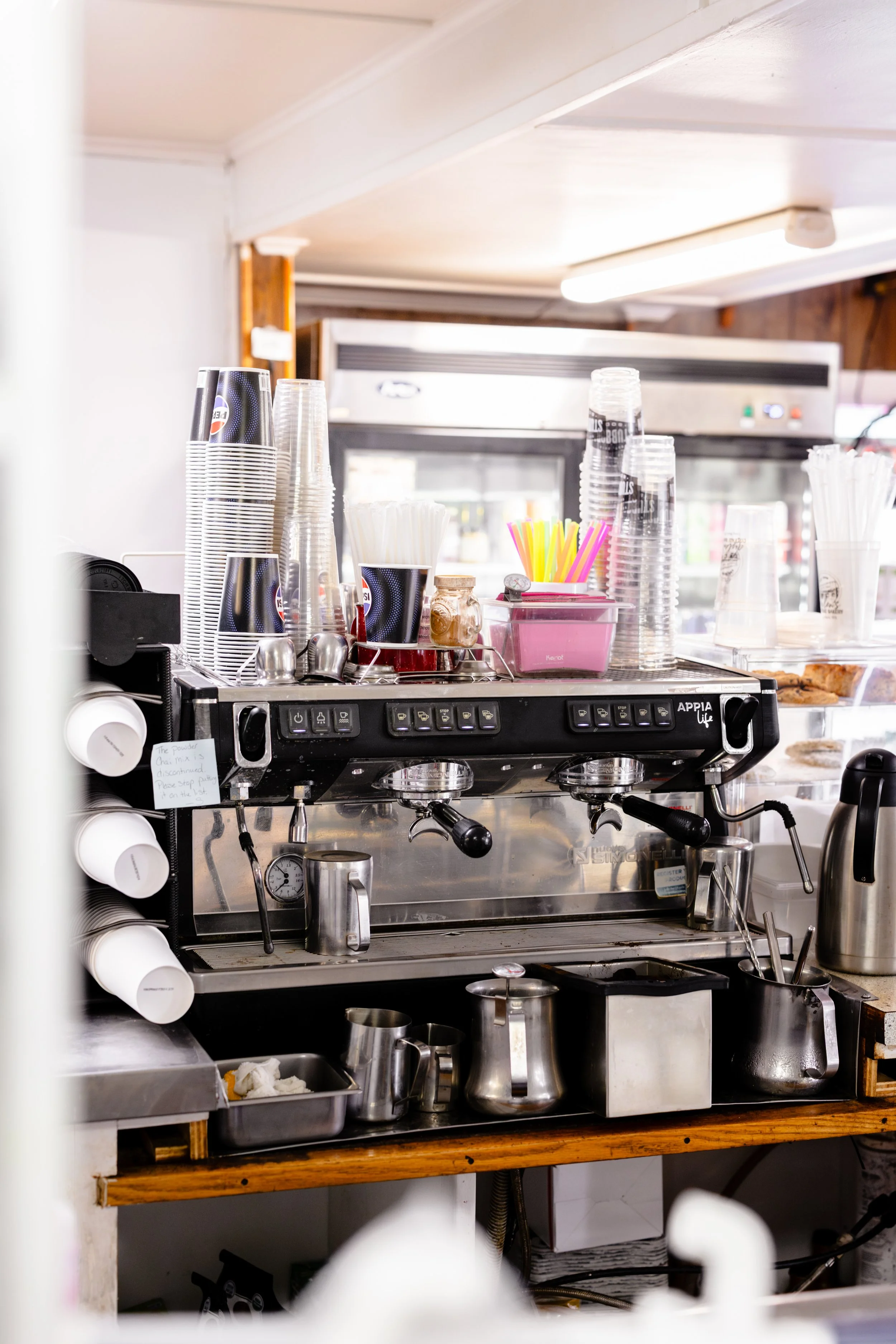 Coffee shop espresso machine with cups, stir sticks, and condiments on top, surrounded by shelves with cups and utensils.