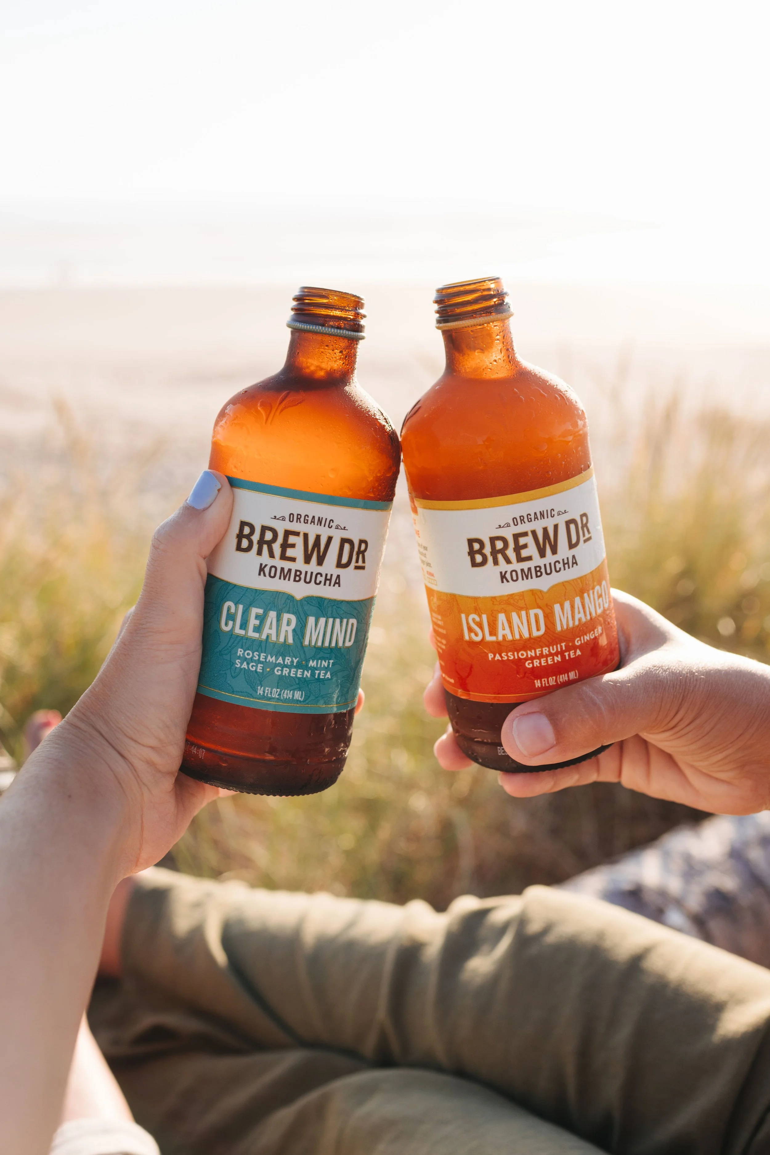 Two people holding bottles of Brew Dr Kombucha, one labeled 'Clear Mind' and the other 'Island Mango,' outdoors near a beach at sunset.