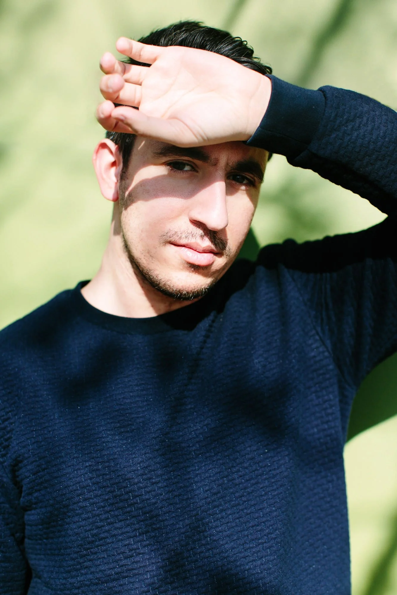 A young man with dark hair and light skin, wearing a navy blue sweater, stands outdoors with a blurred green background. He uses his right arm to shield his eyes from the sunlight, creating a shadow across his face.
